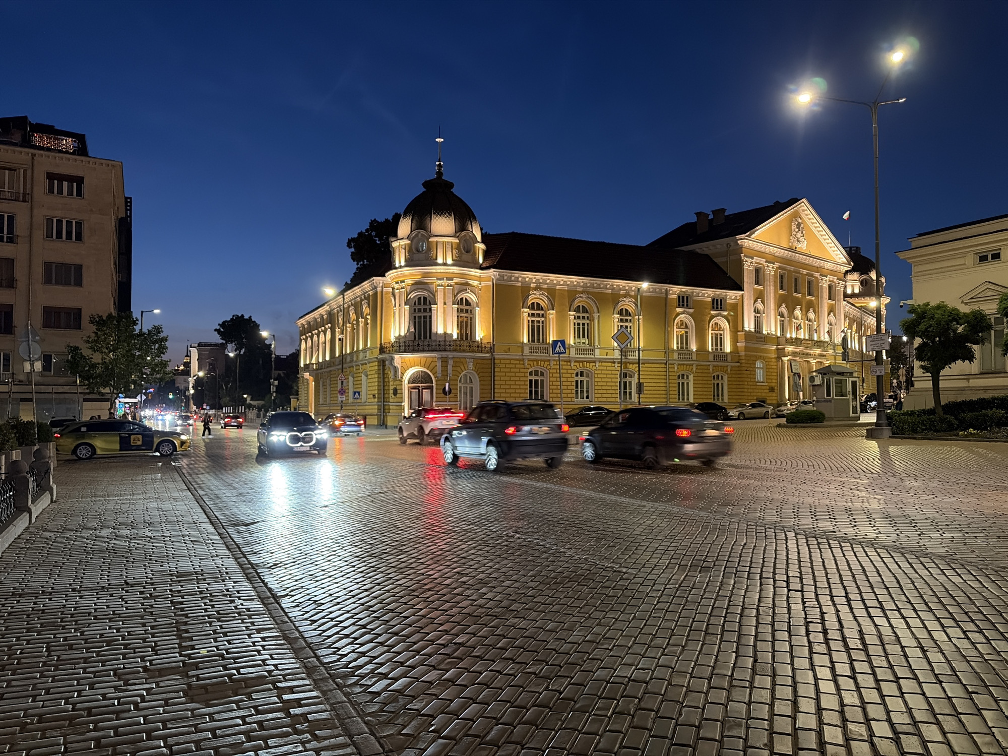 The image depicts a nighttime urban scene in Sofia, Bulgaria, specifically showcasing the former Royal Palace (now the National Art Gallery and National Ethnographic Museum) at Knyaz Alexander I Square.
The scene is dominated by a brightly illuminated, multi-story yellow building on the right, featuring an ornate facade, numerous arched windows, balconies, and a distinctive corner dome topped with a spire. The building is lit with warm yellow artificial lights. To the left of the main yellow building, a taller, less ornate residential or commercial building with rectangular windows is visible. The sky above is a deep, clear blue, characteristic of civil twilight.
The foreground is a wide cobblestone street and pavement, reflecting the artificial light sources. Several vehicles are present on the street, many exhibiting motion blur due to movement. These include a yellow and black taxi-like car parked on the left, and multiple other passenger vehicles (SUVs, sedans) with active headlights and taillights. One vehicle in the mid-ground on the left shows bright white headlights, while several cars further down the road show red taillights. A single pedestrian figure is discernible in the distance, crossing the street.
Streetlights are visible throughout the scene, with a prominent tall streetlight on the right, displaying bright lens flares. Various street signs, including a blue square sign with a white pedestrian symbol, are present near the main yellow building. Decorative metal railings line the left side of the cobblestone foreground. The overall impression is an active city environment during the evening.