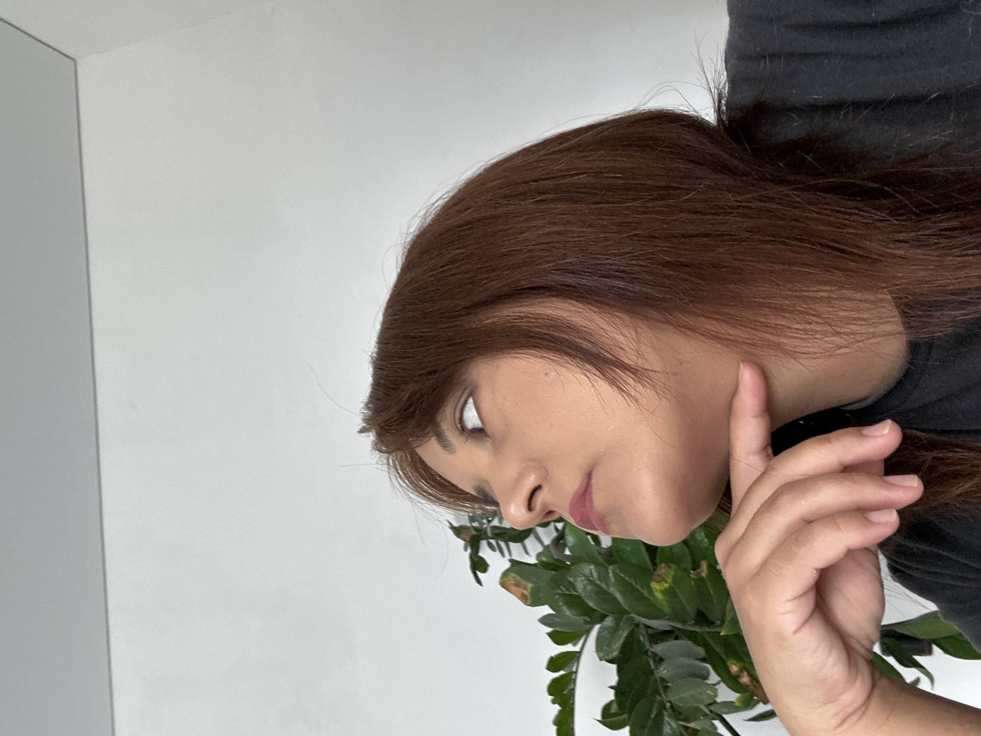 A woman with brown hair is looking downwards, with her chin resting on her hand. There is a green plant visible below her face. The background is a white wall. The location is Unknown, Unknown.