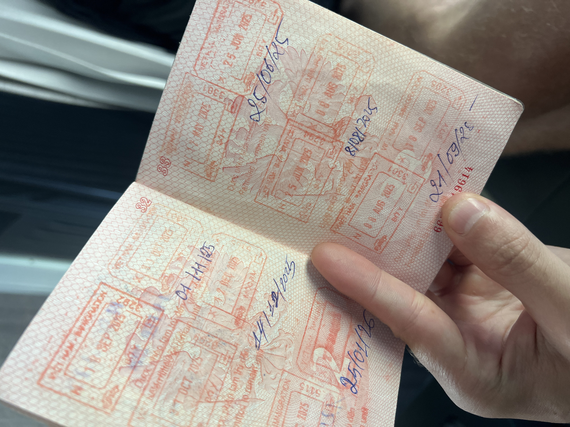 A person is holding open a passport, displaying several visa stamps. The passport pages are printed with a fine, pink, patterned background. The stamps are primarily red ink and feature an ornate design, with specific dates and abbreviations over them written in blue ink. The dates visible on the stamps are: 01.11.125, 14/12/2025, 25/01/26, 25 JUN 2025, 25/06/25, 8/08/2025, and 21/09/23. The stamps include some information related to immigration. Text in the background is "Duoc Nico lam to". The stamps feature a location within Vietnam; Kon Ra Kiah is not explicitly stated in the stamp details.