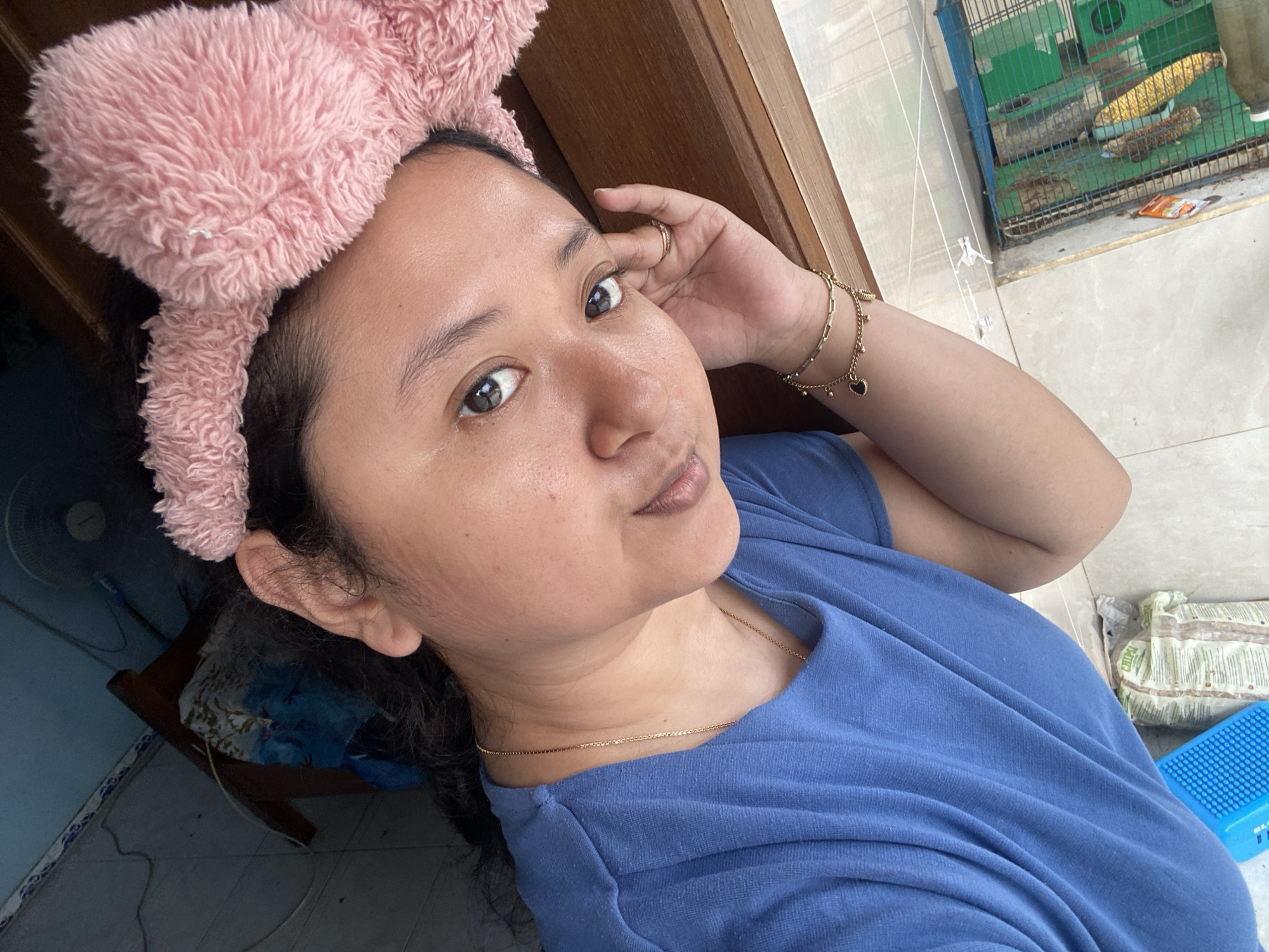 This media file is a high-angle selfie of a woman, likely taken during the daytime in a casual, domestic setting in Gelam, Indonesia. The woman, who appears to be of Asian descent, is the central subject, looking directly at the camera with a relaxed expression and a slight pout. She wears a fluffy, light pink headband styled like bunny ears, pushing back her dark hair. She is dressed in a plain blue t-shirt and accessorized with a thin gold chain necklace and a gold bracelet adorned with small black heart charms. A gold ring is visible on her left index finger, as her left hand rests near her head.
The background details offer a glimpse into her surroundings. To the right of the woman, a green and blue wire birdcage is visible, containing what appears to be corn cobs or other feed. Below the cage, a light-colored tiled floor shows some visible dirt and debris, and a small orange packet with the brand name "CHIPIO" is on the ground. Above the birdcage, white strings with clothesline clips are strung across the space. To the left, a blue electric fan and a piece of wooden furniture are partially visible in a more shaded area. Near her right shoulder, a blue object with the text "SLIM BLU" is also seen on the floor. The overall ambiance is unposed and homey.