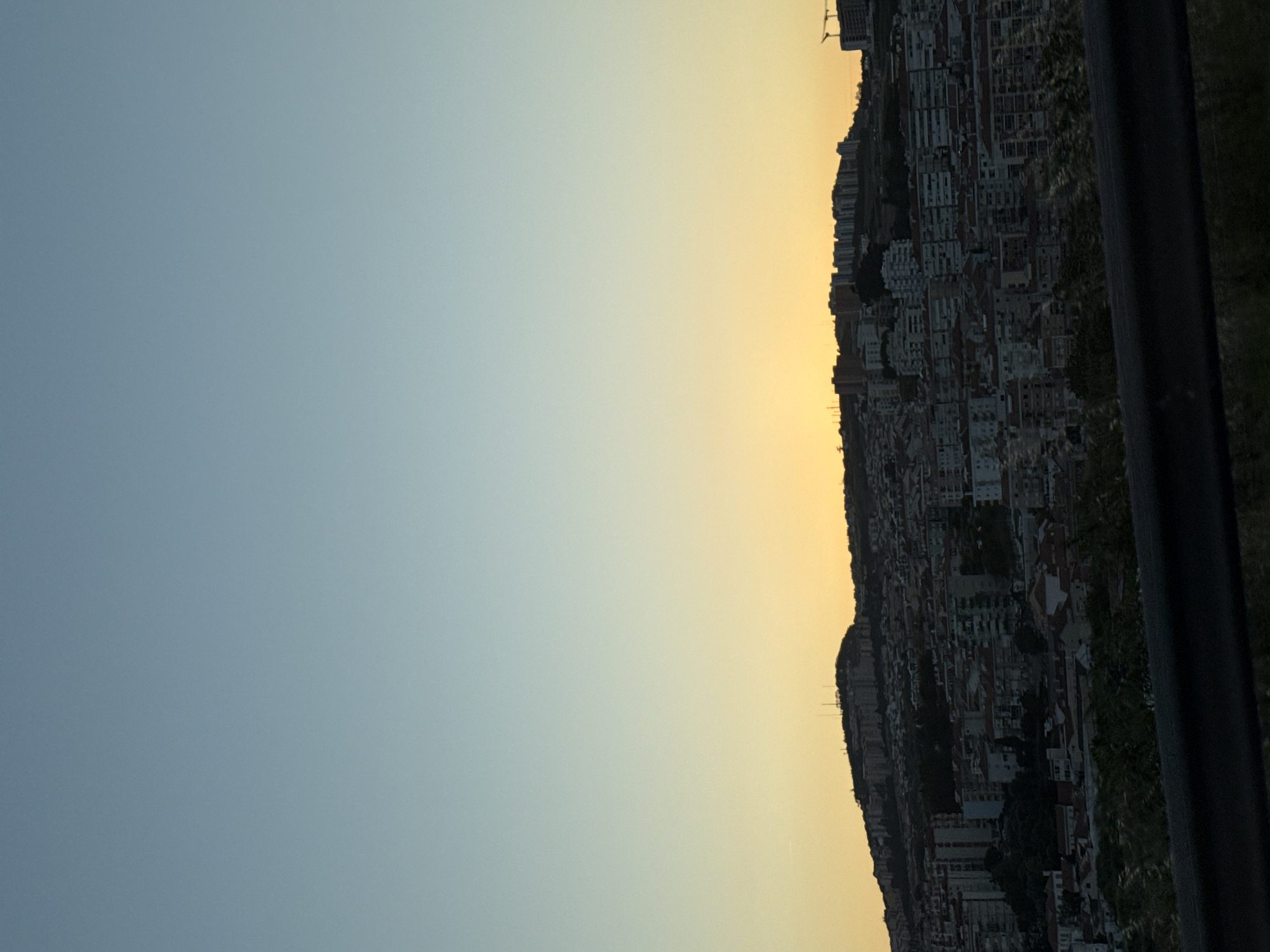 This is a vertical, eye-level, full shot photograph of the city of Loures, Portugal, taken at dusk. The sky is a gradient of clear blue at the top, transitioning to a soft yellow near the horizon where the sun has set. The city is built on a sloping hill, densely packed with buildings of various sizes and styles, predominantly apartment buildings with tiled roofs. Some taller, modern buildings are visible on the crest of the hill, along with communication towers. The foreground features a dark, blurred railing and some indistinct foliage, suggesting the photo was taken from an elevated vantage point, possibly a road or bridge. There are no people visible in the image. The overall scene depicts a sprawling urban landscape under a serene evening sky.