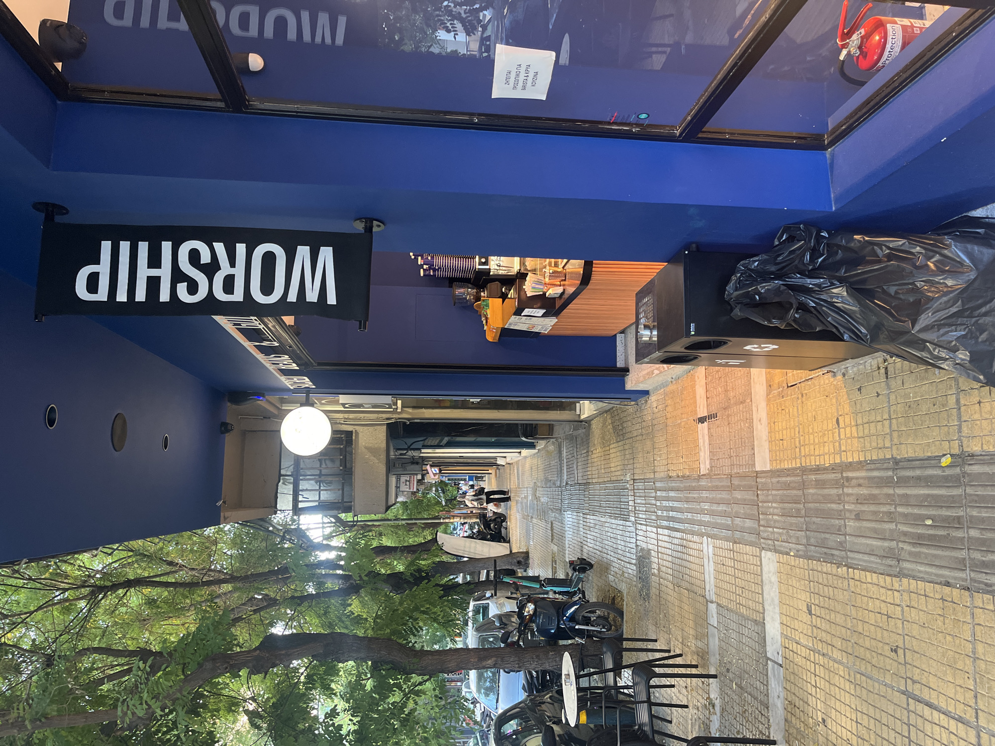 The image shows a street-level view of a coffee shop in Athens, Greece. The shop has a bright blue facade with a large window and a prominent black banner with white lettering that reads "WORSHIP." Inside the shop, a wooden counter displays coffee-making equipment and cups. On the sidewalk outside, there are chairs and tables, a trash can with a black garbage bag, and several parked scooters and motorcycles. A fire extinguisher is visible near the shop's entrance. The sidewalk is paved with yellow tiles and has a tactile paving strip for visually impaired pedestrians. Trees line the street, providing shade. A few people are walking on the sidewalk in the background. A sign on the window is written in Greek and appears to be a job advertisement.