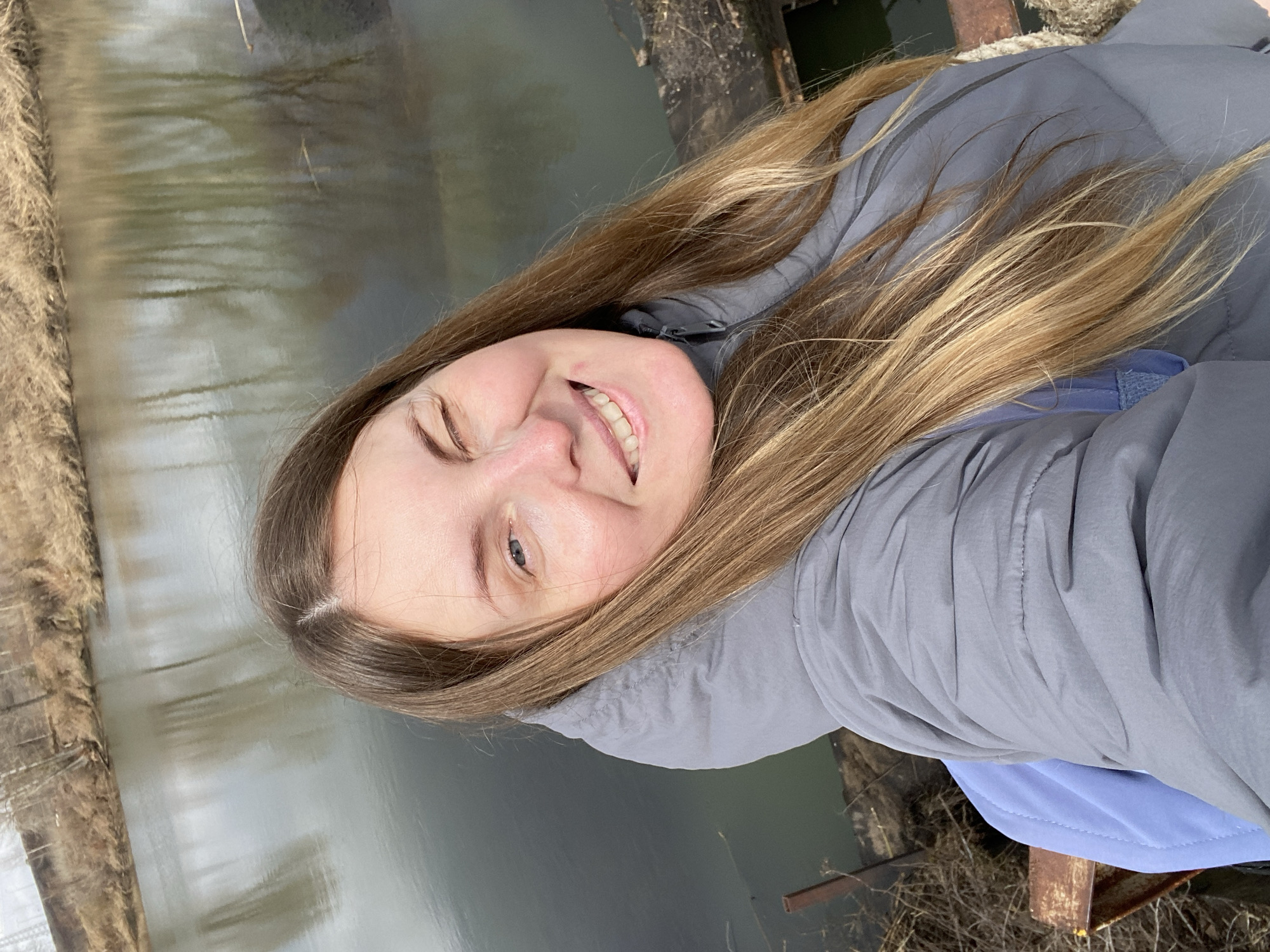 A woman is taking a selfie outdoors. She is smiling with one eye closed and has long, light brown hair. She is wearing a gray jacket with a blue underlayer. Behind her, there's a body of water reflecting the sky and some kind of wooden or metal structure with dried vegetation. The location is unspecified.