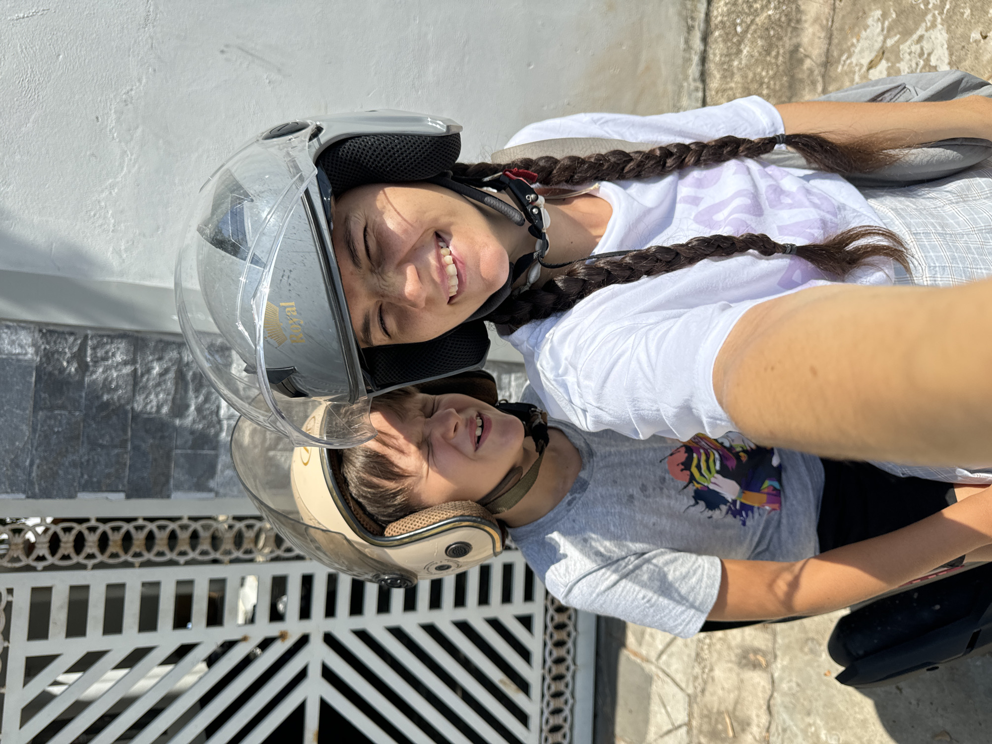 In Mũi Né, Vietnam, two individuals, a woman and a child, are captured in a selfie on a sunny day. The woman, in the foreground, wears a white t-shirt and a gray plaid skirt, her dark hair styled in two braids. She has a silver helmet with a transparent visor, and a bright smile, her eyes squinted against the sun. Next to her, the child, wearing a gray t-shirt, sports a tan helmet with a clear visor, also squinting and smiling. Both are seated on what appears to be a scooter, with the woman holding the camera. Behind them, a white metal gate with a geometric pattern is visible, along with a textured gray wall, suggesting an outdoor urban setting. The ground is paved with rough concrete tiles.
