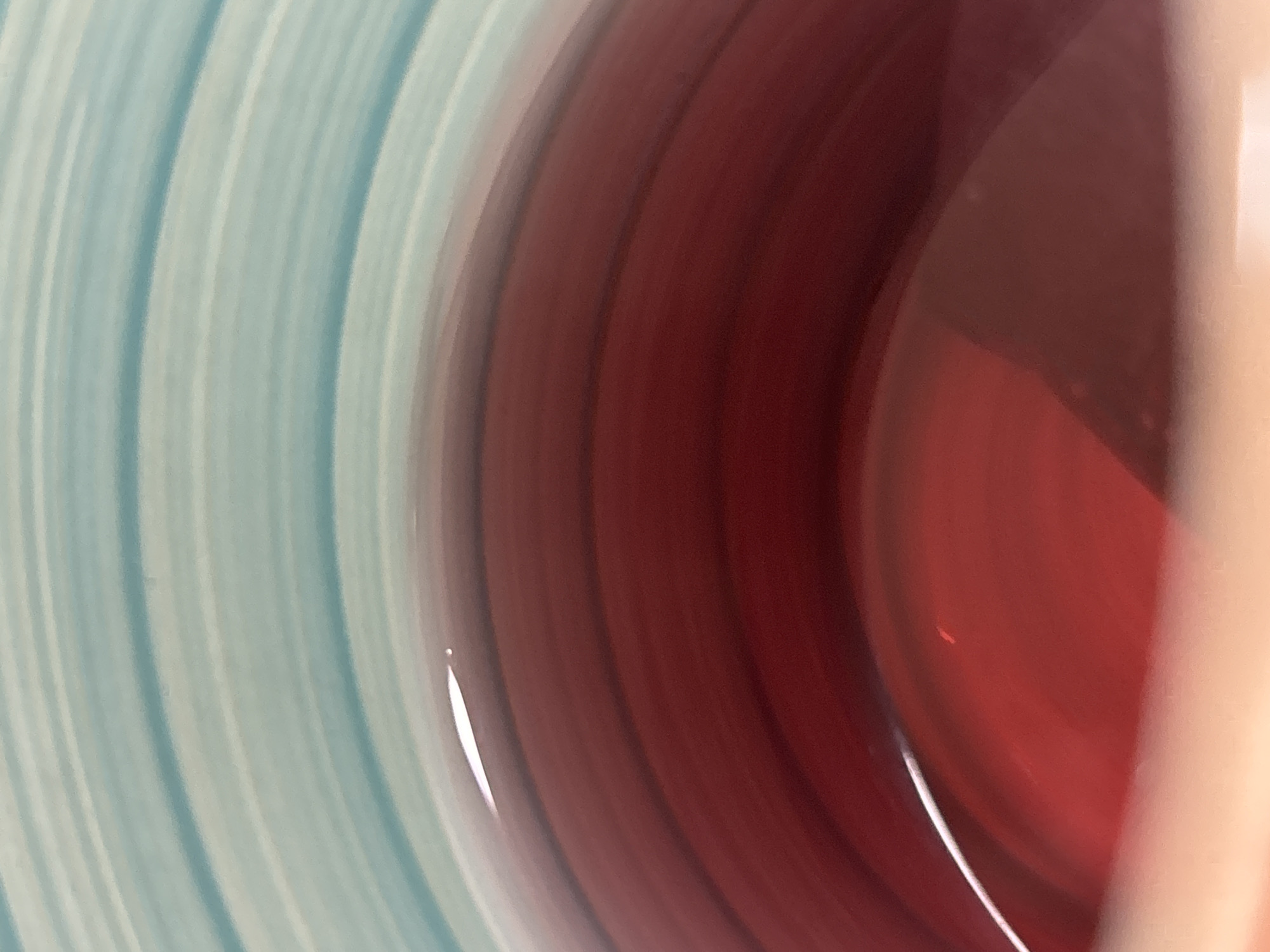 The image is a close-up, abstract shot of the interior of a mug or cup. The upper portion of the image features horizontal stripes of light blue and white, suggesting the pattern of the mug's exterior. Below this, the dominant feature is a pool of deep red liquid, likely a beverage, filling the lower part of the mug. The liquid's surface is smooth and slightly curved, reflecting the light with a bright highlight along its edge. Subtle darker red bands are visible within the liquid, possibly indicating depth or variations in density. The bottom of the mug appears to be a pale orange or peach color, visible at the very bottom edge of the frame. The image does not contain any elements that would allow for identification of a specific location in the city of Ostrov, Russia.