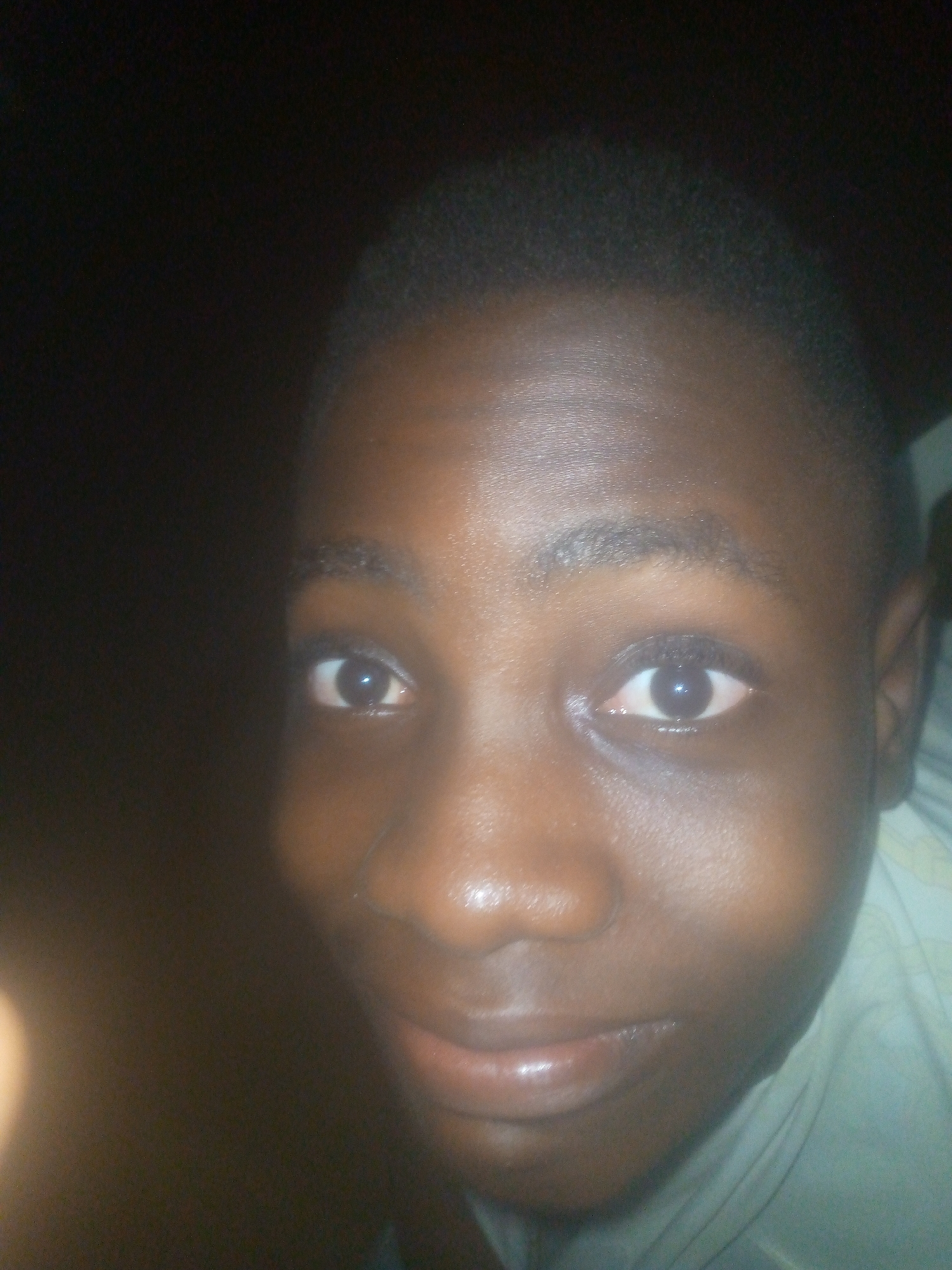 A close-up headshot features a young individual with dark skin, looking directly at the camera. The person's face is illuminated by a direct light source, likely a camera flash, which creates prominent reflections on their forehead, nose, and lips, as well as distinct catchlights in their wide-open dark eyes. Their expression is neutral with a slight hint of a smile. Short, dark, and curly hair is visible on the top of their head. A light-colored garment, possibly a shirt, is visible on their right side. The background is almost entirely dark and indistinct, offering no specific location details. The dim ambient lighting suggests the photograph was taken in a very low-light environment, such as indoors at night. No text is visible in the image.
