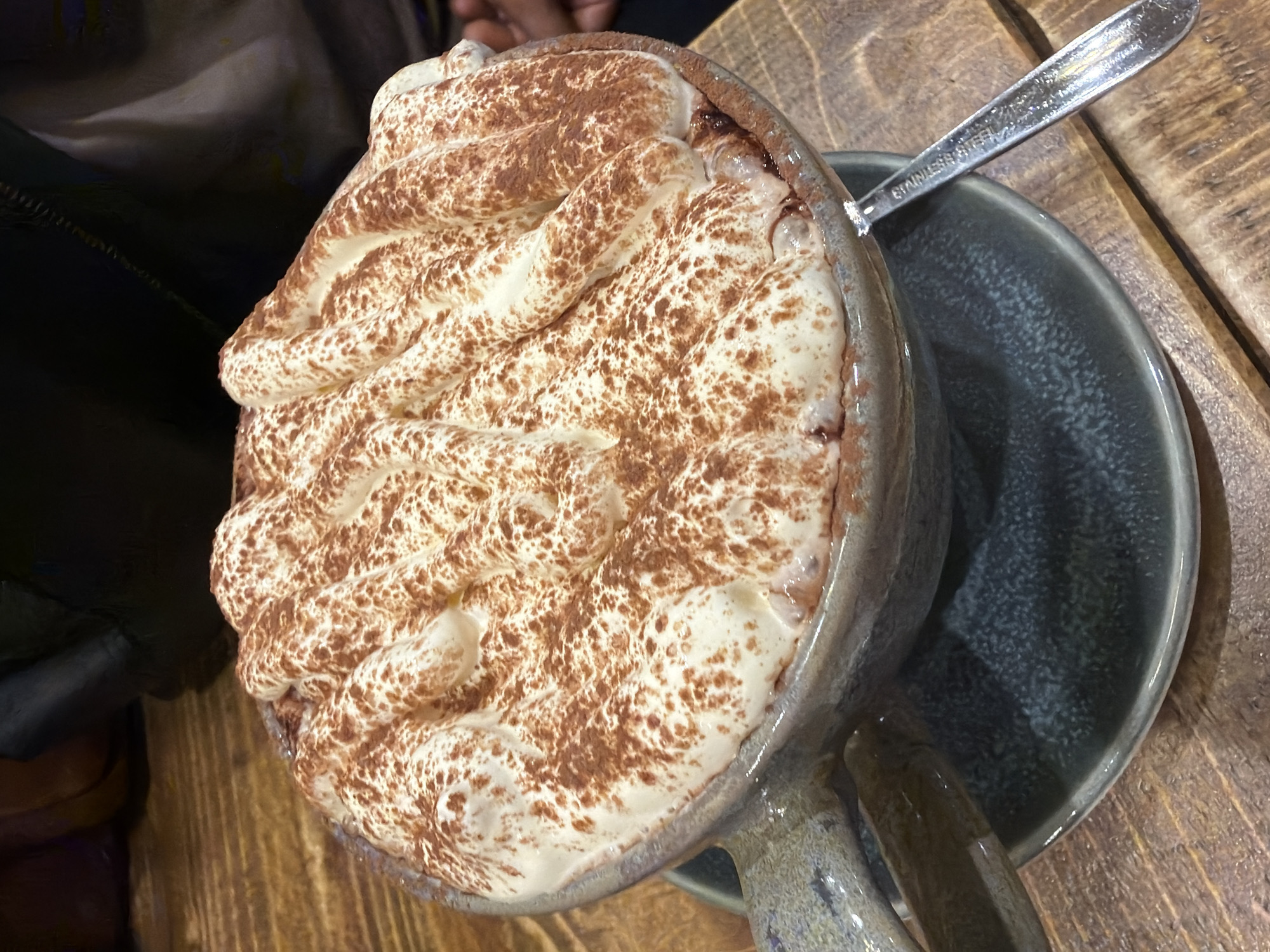A close-up, angled shot shows a mug of hot chocolate topped with whipped cream and dusted with cocoa powder. The mug is a light blue-gray ceramic and sits on a matching saucer on a wooden table. A stainless steel spoon rests partially on the saucer and mug. A person's shoulder and arm are visible in the upper left, suggesting the beverage is being enjoyed at a table. The location within Mole Valley, United Kingdom cannot be determined from this image.
