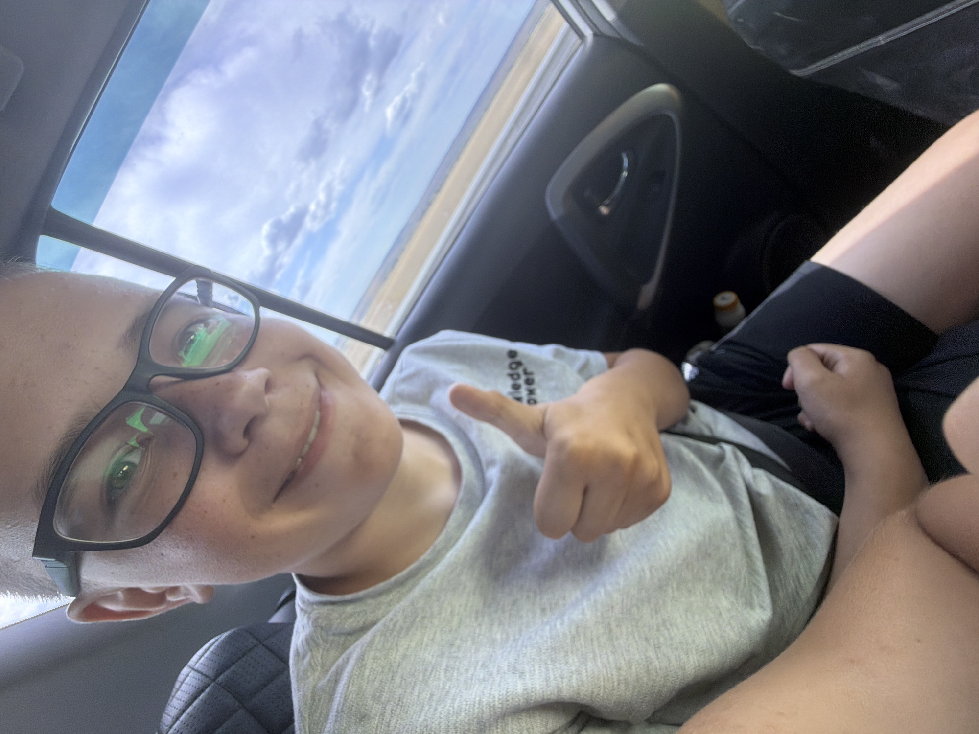 In this media file, a young person wearing glasses and a grey t-shirt is captured in what appears to be a car. The person is smiling and giving a thumbs-up gesture, suggesting a positive emotion and possibly a sense of approval or well-being. The background shows the sky with clouds and what looks like a flat landscape, possibly indicative of a road trip or travel. The interior of the car is visible, including a car door handle and a seat, suggesting the setting is inside a vehicle. The time of day seems to be daytime, given the brightness of the sky.

There is visible text on the t-shirt, though it is partially obscured and difficult to read in full. The visible words appear to be "knowledge is power." No other significant objects or actions are clearly discernible beyond the person and their immediate surroundings. The overall impression is a casual, personal snapshot taken during a journey.