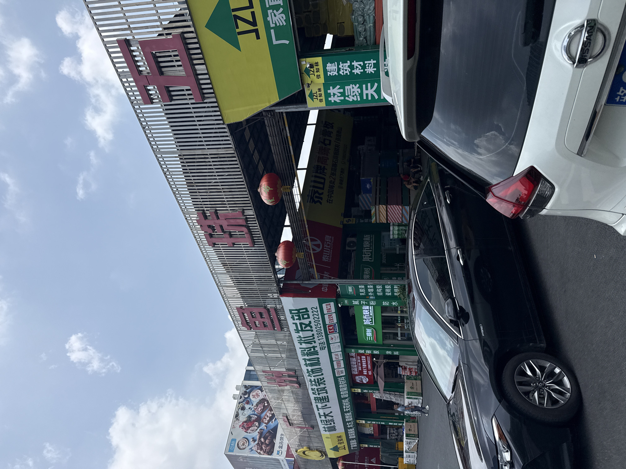 The image captures a commercial street scene in Tianhe District, China, during bright daytime with a partly cloudy sky. In the foreground, two vehicles are parked: a black sedan, displaying "HYBRID" on its side, and a white Nissan SUV, identifiable by its rear "NISSAN" badge.
The left side of the street is lined with shops specializing in building and decoration materials, featuring large, multi-colored signs and awnings. A red Chinese lantern hangs from one of the overhead structures. A few people are faintly visible near the shops.
Numerous signs display Chinese text, advertising various businesses and products. Notable visible text includes:
* **Shop names**: "赢绿天下建筑装饰材料批发部" (Yinglu Tianxia Building Decoration Materials Wholesale Department), "林绿天下建材批发部" (Linlu Tianxia Building Materials Wholesale Department), "JZL 佳知林" (JZL Jiazhilin), "超级" (Super), and "盒马鲜生" (Hema Fresh - potentially an advertisement or nearby store).
* **Products/Brands**: "泰山牌局端石膏板" (Taishan Brand Gypsum Board), "三棵树" (Three Trees) for "乳胶漆" (emulsion paint), "防水" (waterproof materials), "涂料" (paint), and "结构胶" (structural adhesive). Other products mentioned are "建筑材料" (building materials), "德高瓷砖胶" (Degao Tile Adhesive), "建筑模板" (construction formwork), "木方" (wood), and "阻燃板" (fire-retardant boards).
* **Slogans/Details**: "厂家直" (Factory Direct), "城市焕新" (Urban Renewal).
* **Contact information**: Phone numbers like "13802502222" and "139602222".
The scene suggests an active commercial area dedicated to construction and home improvement supplies.