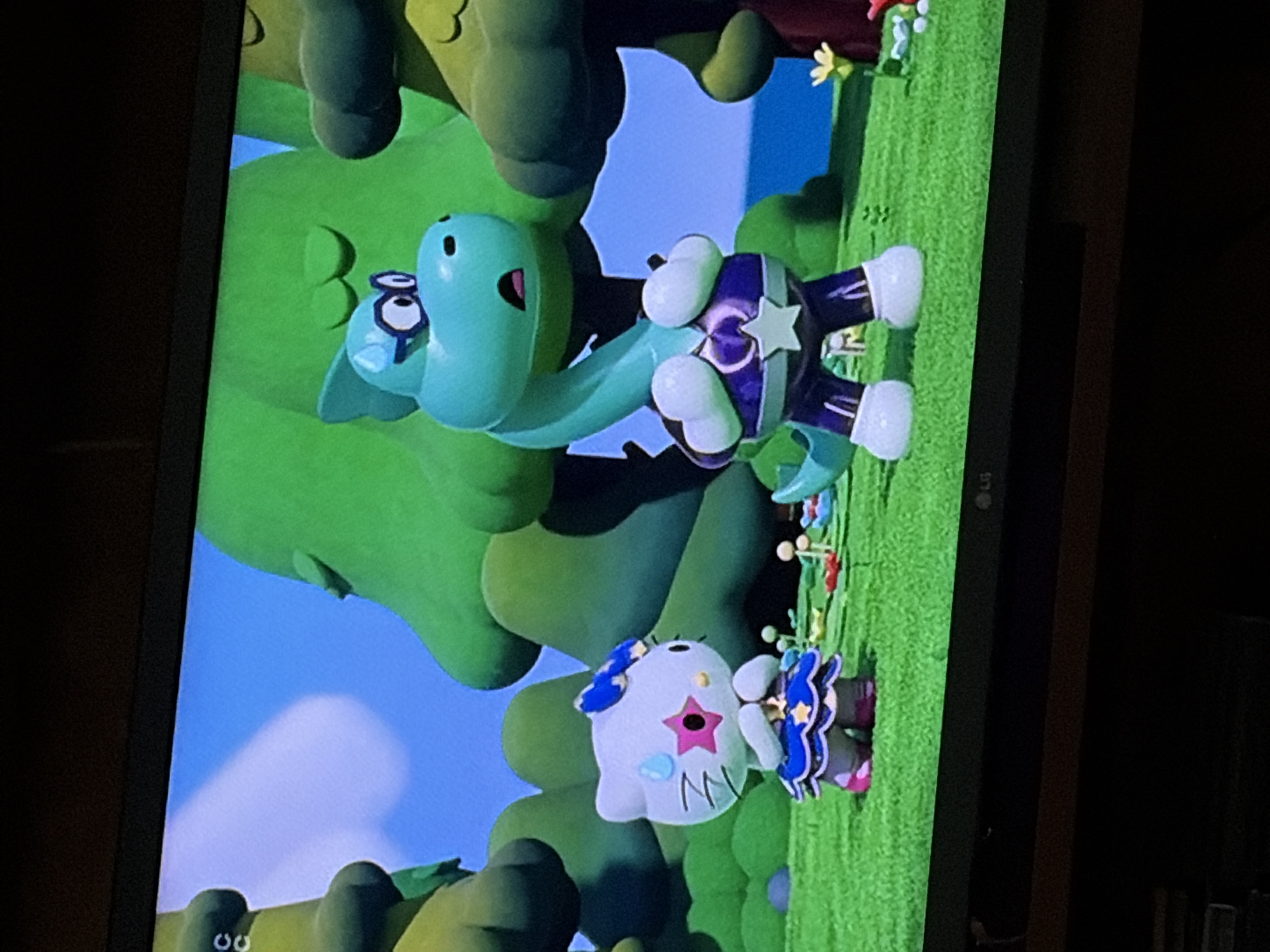 In a brightly lit outdoor scene with a blue sky and green grass, two animated characters stand in a grassy clearing. On the left, a white anthropomorphic cat with a blue bow on its head wears a blue and gold dress. The cat has a pink star with a smaller star inside on its cheek and a sweat drop above its head. It is holding its hands together in front of its chest. On the right, a turquoise dinosaur wearing a purple and white costume with a star emblem stands upright. The dinosaur has large black-rimmed glasses and an open mouth, appearing surprised. Behind the characters are large, stylized green trees. The scene is consistent with a digital animation or a television screen display. The background includes hints of smaller plants and flowers scattered on the grass.