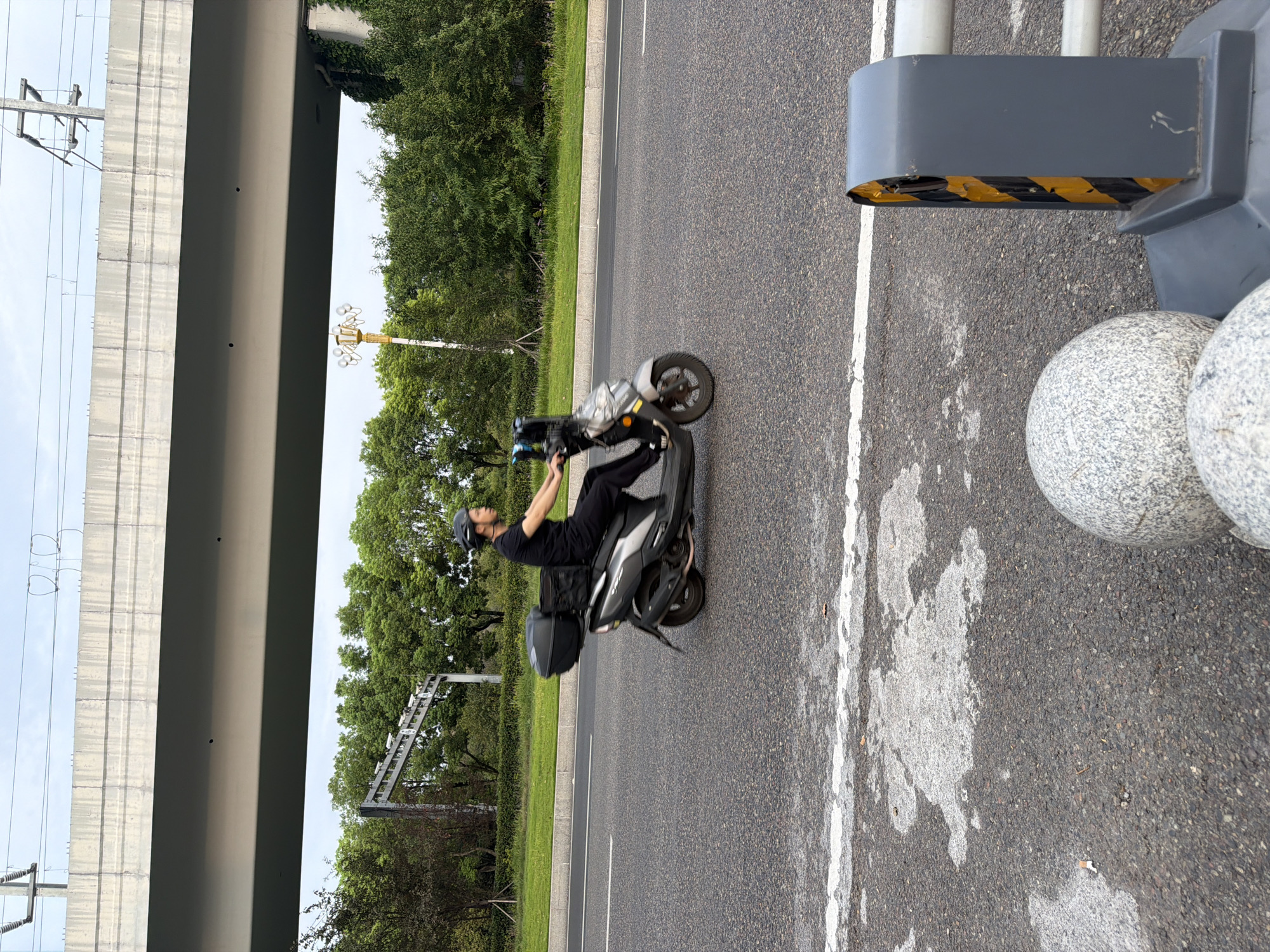 A person wearing a helmet and a black t-shirt and pants rides a silver scooter on a paved road. The scooter has a black storage box mounted on the back. The road is lined with a white stripe. In the background, there are trees and a large elevated concrete structure. The sky is overcast. The scene is in Jiangjin, China.