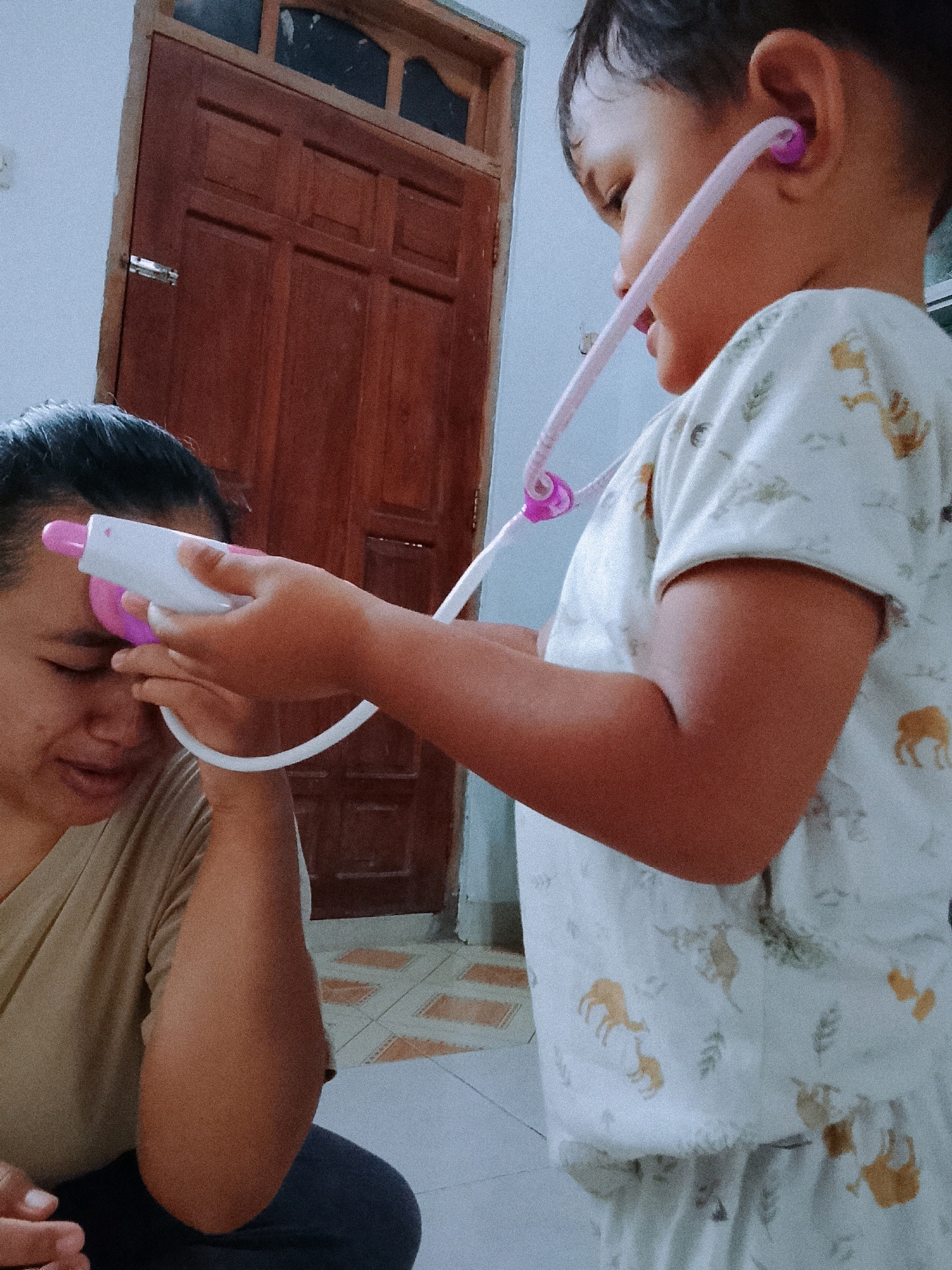 In a room in Kradenan, Indonesia, a young child is playing doctor with an adult. The child, wearing pajamas with a deer pattern, holds a pink toy stethoscope to the adult's forehead. The adult, with dark hair pulled back, wears a tan shirt and has their eyes closed, seemingly participating in the game. A wooden door with metal hardware is visible in the background, along with tiled flooring.