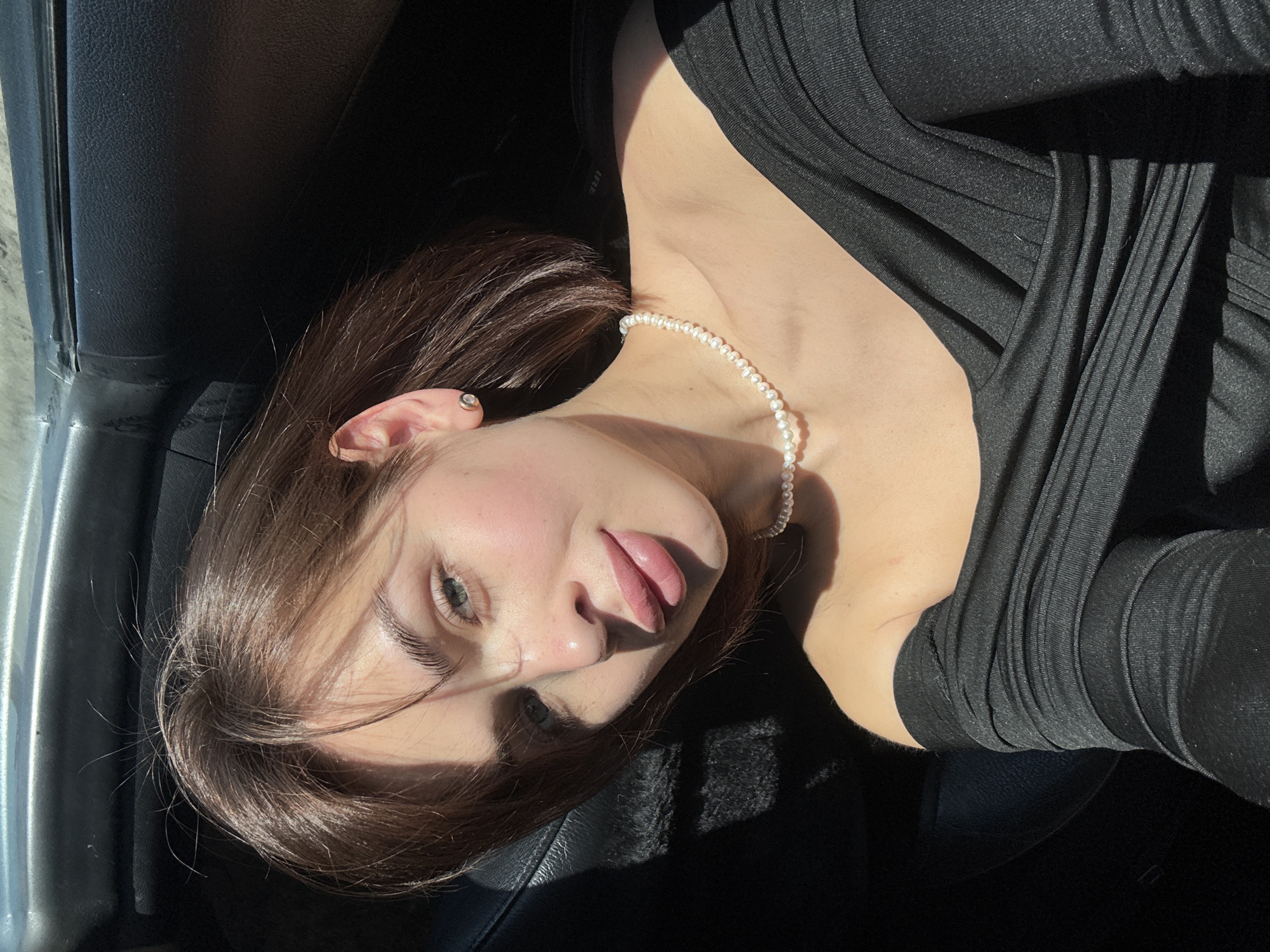 A young woman is captured in a close-up, low-angle shot inside a vehicle. She is turned to the side, facing the camera with her lips slightly parted. She is wearing a dark, long-sleeved shirt and a pearl necklace. Part of a car door is visible in the upper left of the frame. The location is Лобаново, Russia.