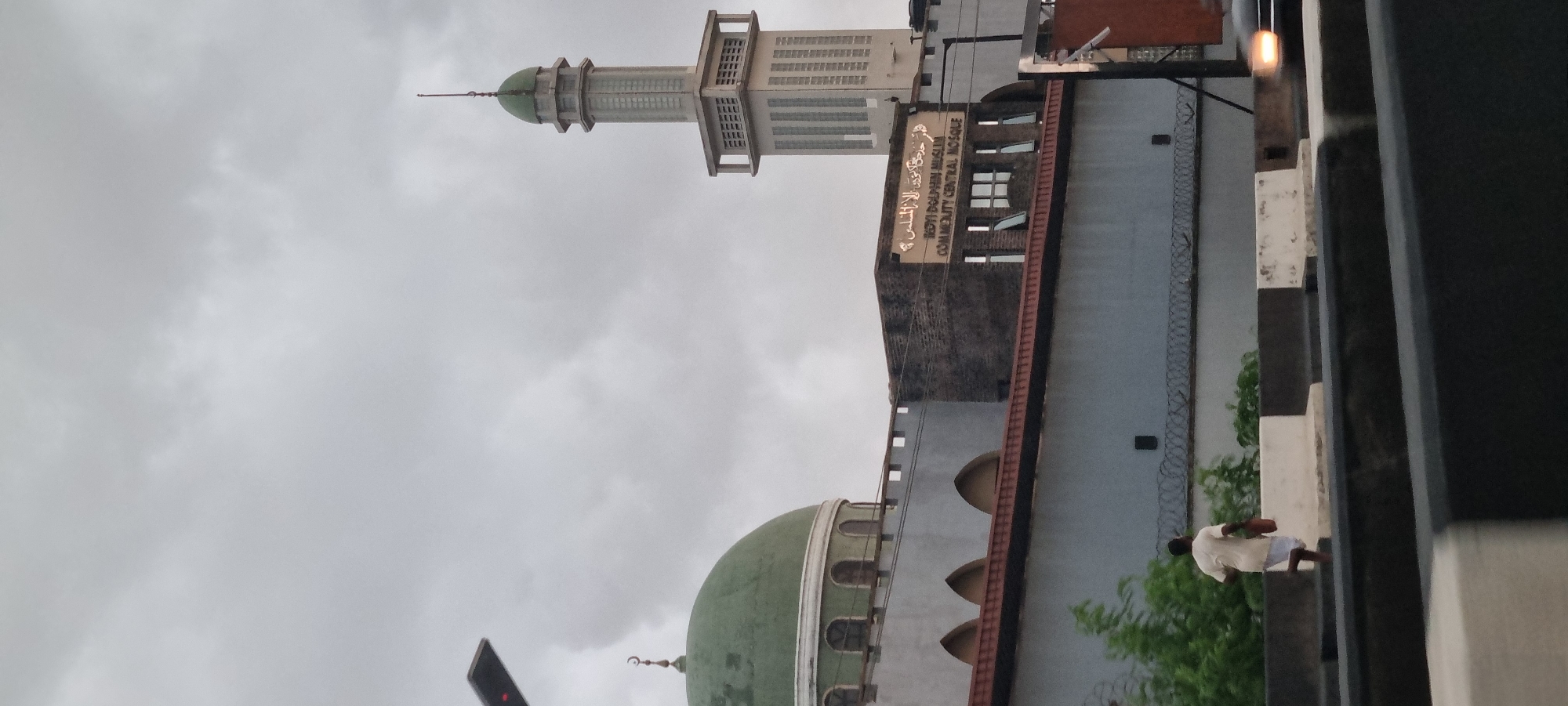 The image shows the external view of the Ikoyi Dolphin Muslim Community Central Mosque in Ikoyi, Nigeria. The mosque features a large green dome and a tall minaret with a green cap. A sign on the building reads "IKOYI DOLPHIN MUSLIM COMMUNITY CENTRAL MOSQUE" in English, with Arabic script above it. A person in a light-colored robe and white shorts is walking away from the camera, carrying a red bucket. The foreground shows a road with traffic barriers, and the sky is overcast.