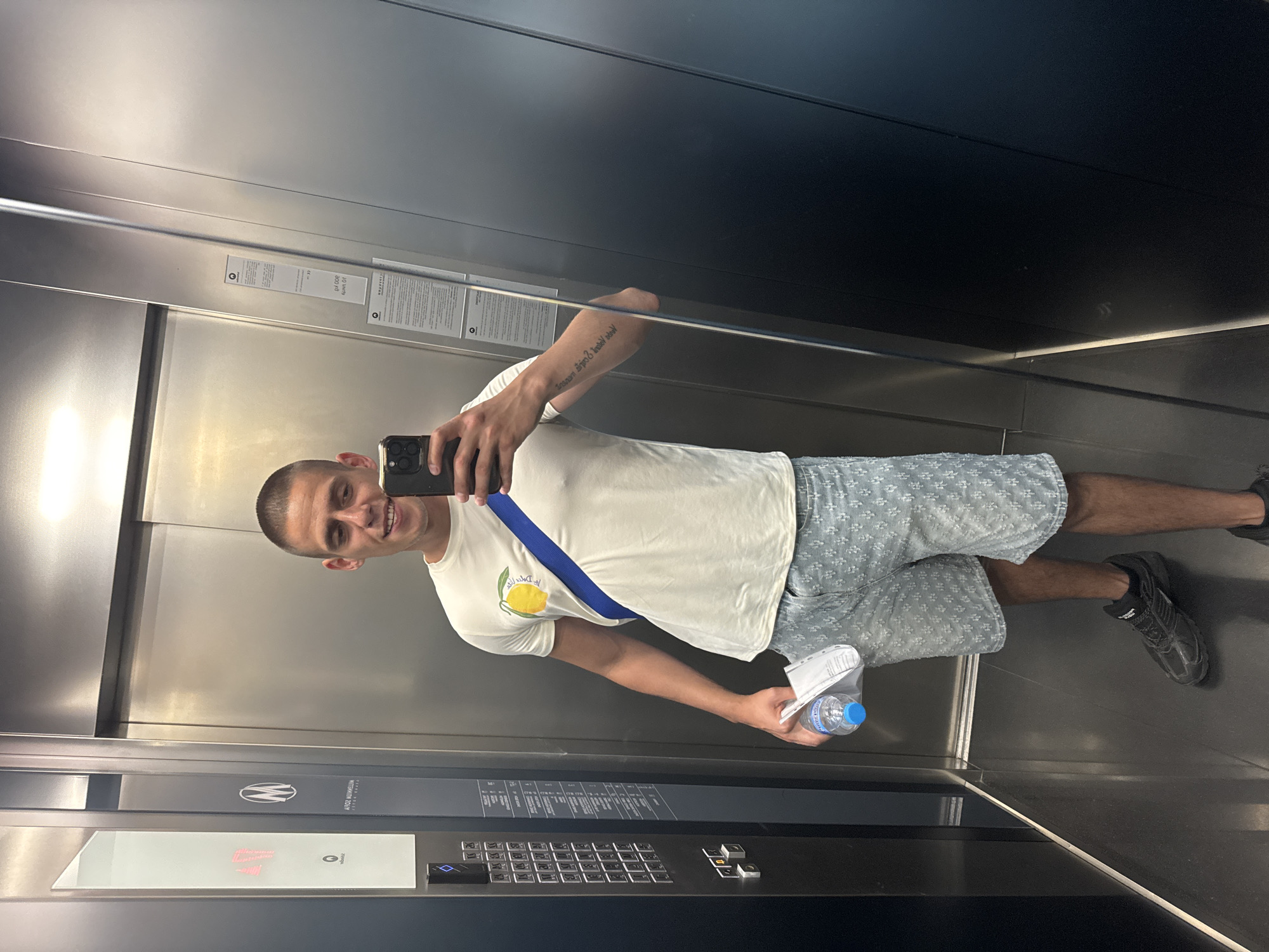 A man is taking a selfie in an elevator. He is wearing a white t-shirt with a lemon graphic, patterned blue shorts, and black shoes. He is holding a phone in one hand and a water bottle in the other. The elevator has metallic walls and a control panel with buttons. The elevator display reads "14" suggesting he is on the 14th floor. The elevator has an "Anton Montpellian" logo. The location is Sofia, Bulgaria.