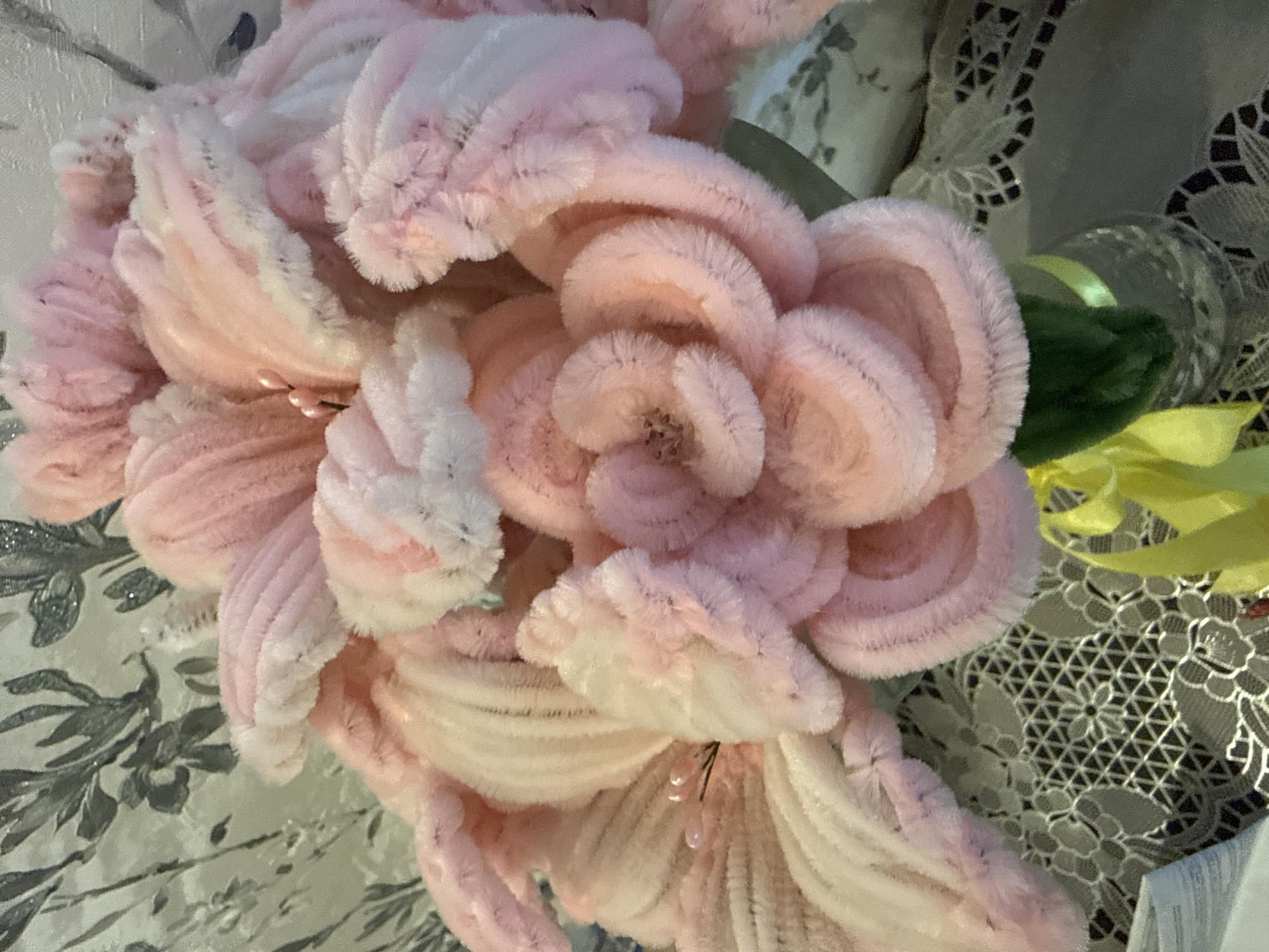 The image shows a close-up of artificial pink and white flowers, possibly made from chenille stems. Some green and yellow ribbons are visible behind the flowers. A patterned, lace-like surface can be seen in the background. The location in the city of Марковский, Russia is not evident from the image.