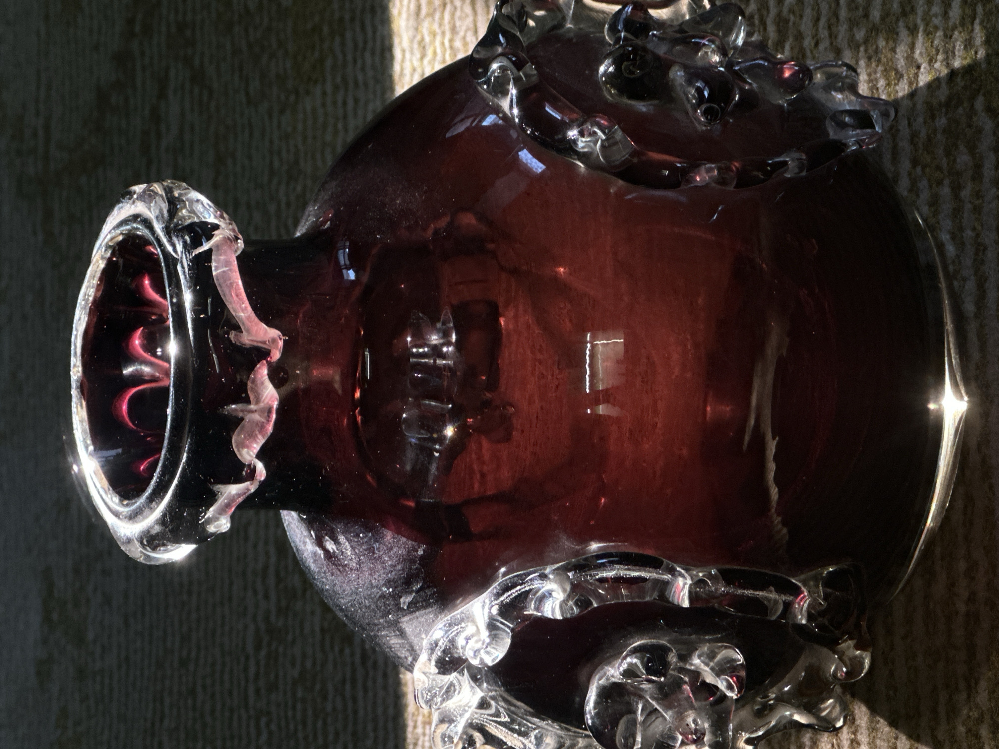 A close-up, overhead shot shows a dark red glass vase with ornate, clear glass embellishments on its sides and rim, set against a textured, light brown surface. The vase is illuminated by strong, direct sunlight casting sharp shadows. The glass is reflective, showing highlights and blurred reflections of its surroundings. The scene is an indoor setting, and there is no information available to indicate its location in Luhansk, Ukraine.