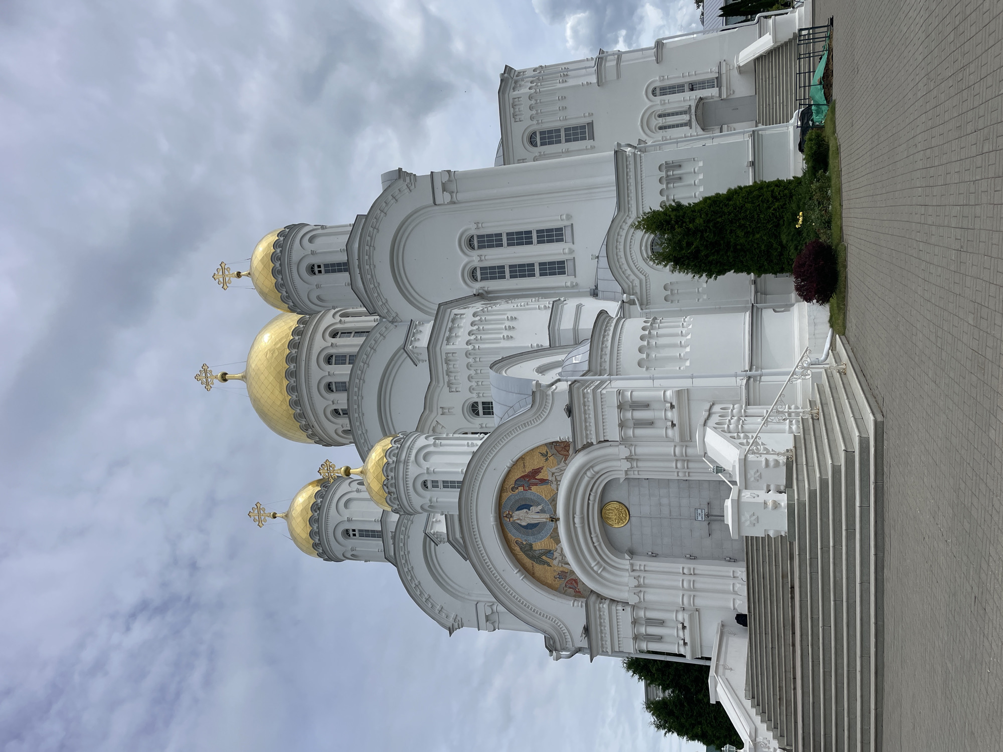 A large, white Orthodox cathedral with golden domes stands under a cloudy sky. The cathedral has multiple domes topped with crosses. The facade features intricate architectural details, including arched windows and decorative moldings. A mosaic depicting a religious scene is situated above the main entrance. A wide set of stone stairs leads up to the entrance, with white railings on either side. Green trees and landscaping are visible in front of the cathedral. The ground in front of the stairs is paved with gray bricks. The scene appears to be a still, exterior view of a religious site. The location is the Seraphim-Diveyevo Monastery in Diveyevo, Russia.