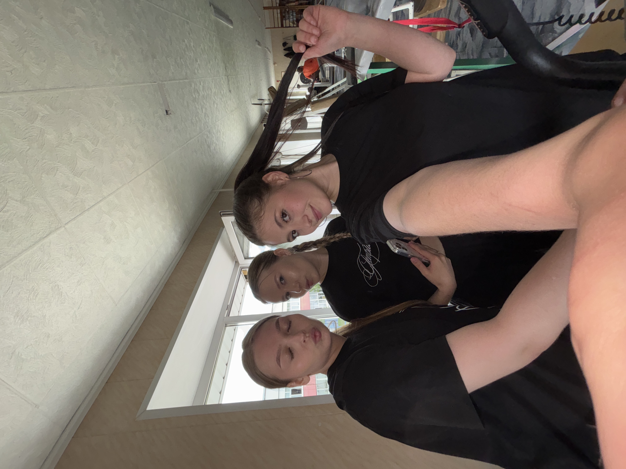 Three young women are taking a selfie in what appears to be a gym or fitness facility in the city of Chaikovsky, Russia. The woman on the left is looking at the camera with her eyes closed and lips pursed. The woman in the middle is holding a phone and looking at the camera with her lips pursed. The woman on the right is holding her long dark hair up and to the side and looking at the camera with a smile. They are all wearing black t-shirts. The background shows gym equipment and a window with a view of buildings. The ceiling is paneled with a textured finish.