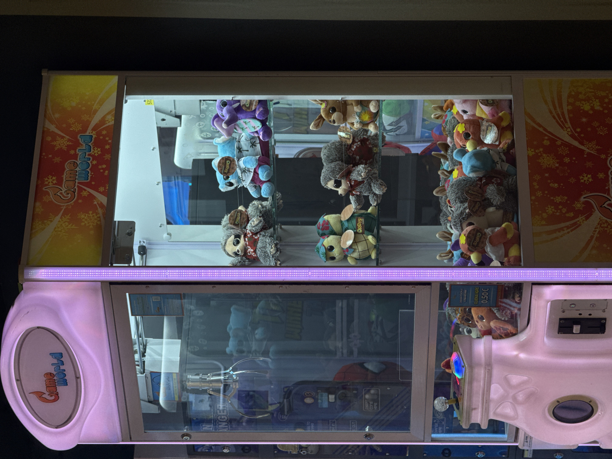 The image shows a claw machine game filled with stuffed animals. The claw machine features a pink and white design with the words "Game World" printed on its side. The stuffed animals inside the machine include various characters, such as sloths, rabbits, and bears, some of which have tags with the word "ORBYS" on them. The machine's interior is lit with a purple light. The text "INSERT COINS HERE" and "CIJENA IGRE 0.50€" can be seen. It is assumed that the location of the claw machine is in an arcade.
