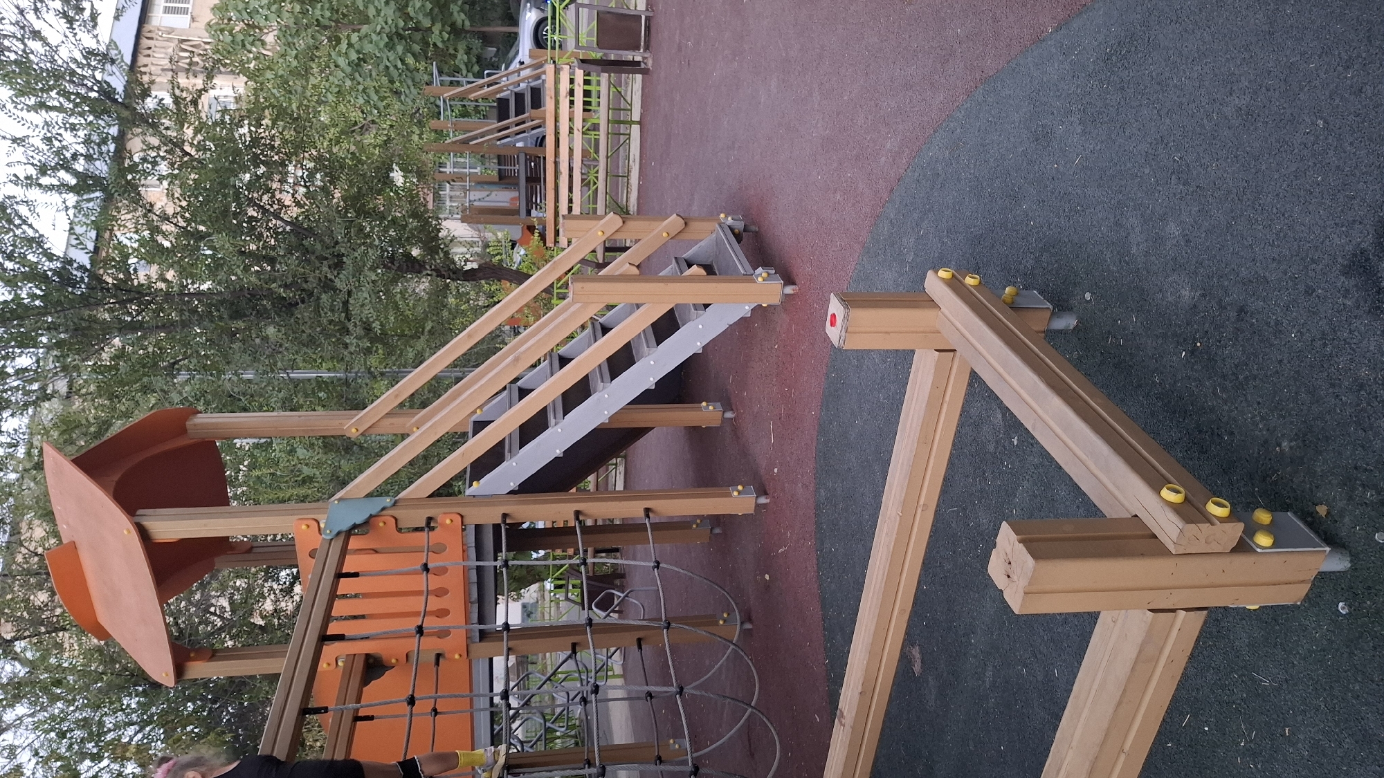 The image depicts a children's playground in an urban setting, specifically in Shymkent, Kazakhstan. In the foreground, a wooden structure with parallel wooden beams supported by vertical posts and metal legs is visible. The beams are secured with yellow caps. The ground beneath is a textured surface of alternating dark gray and reddish-brown rubber.
Further back, a multi-level playground structure is prominent. It features an orange roofed cubby, wooden support posts, and a set of gray stairs with wooden railings. To the left of this structure, a rope net climber is partially visible. In the background, trees with green foliage and a multi-story building can be seen. Beyond the playground, a wooden bench and a metal trash can are situated next to a fence. A silver car is parked to the right of the bench. A person, partially visible on the left side of the image, is wearing black clothing and yellow shoes, and has pink hair tied up. The overall scene suggests an outdoor recreational area within a city.