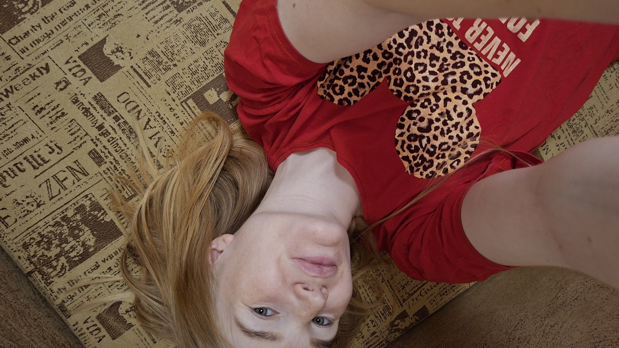 A young woman with fair skin, freckles, and blonde hair lies on a couch covered in a newspaper-print fabric. She is wearing a red t-shirt with a leopard-print design on the chest that resembles Mickey Mouse ears and the text "NEVER FORGET". Her head is tilted back, and she is looking up at the camera with a slight smile. Her arms are raised, with one hand possibly holding the camera. The lighting suggests it is daytime. The room's decor is not visible beyond the couch. The text on the couch fabric includes words like "Weekly", "VIDA", "ZEN", and "MANDO".