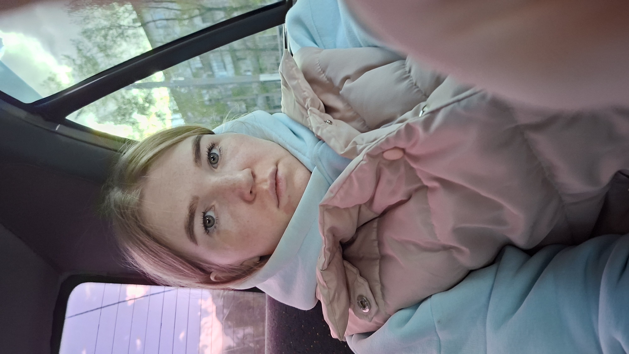 A young woman with blonde hair and fair skin is pictured inside what appears to be a vehicle, potentially a car or a bus. She wears a light blue hoodie and has a beige puffer jacket wrapped around her shoulders. Her face is partially visible, showing her eyes, nose, and lips. The interior of the vehicle is dark, with a window showing a blurry view of trees and buildings, suggesting an urban environment. The location is not verifiable as Chaikovsky, Russia, without further information or context within the image.