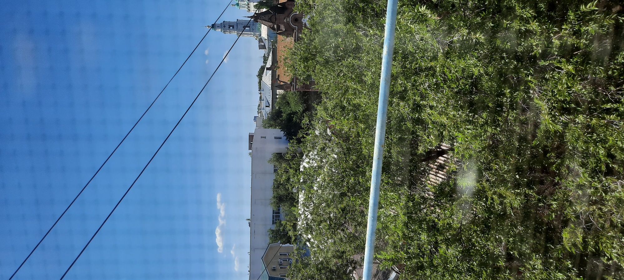 The image depicts a view of the Astrakhan Kremlin, a historic fortress complex located in Astrakhan, Russia. In the foreground, lush green foliage dominates the lower half of the frame, with a light blue metal railing running horizontally across the image. Above the foliage, several buildings are visible, including a white rectangular structure and a traditional house with a green roof. In the background, a prominent white bell tower with a green dome, characteristic of Russian Orthodox churches, rises against a clear blue sky. The bell tower is part of the Assumption Cathedral within the Kremlin complex. The image appears to have been taken through a window, as evidenced by the faint grid pattern superimposed on the scene.