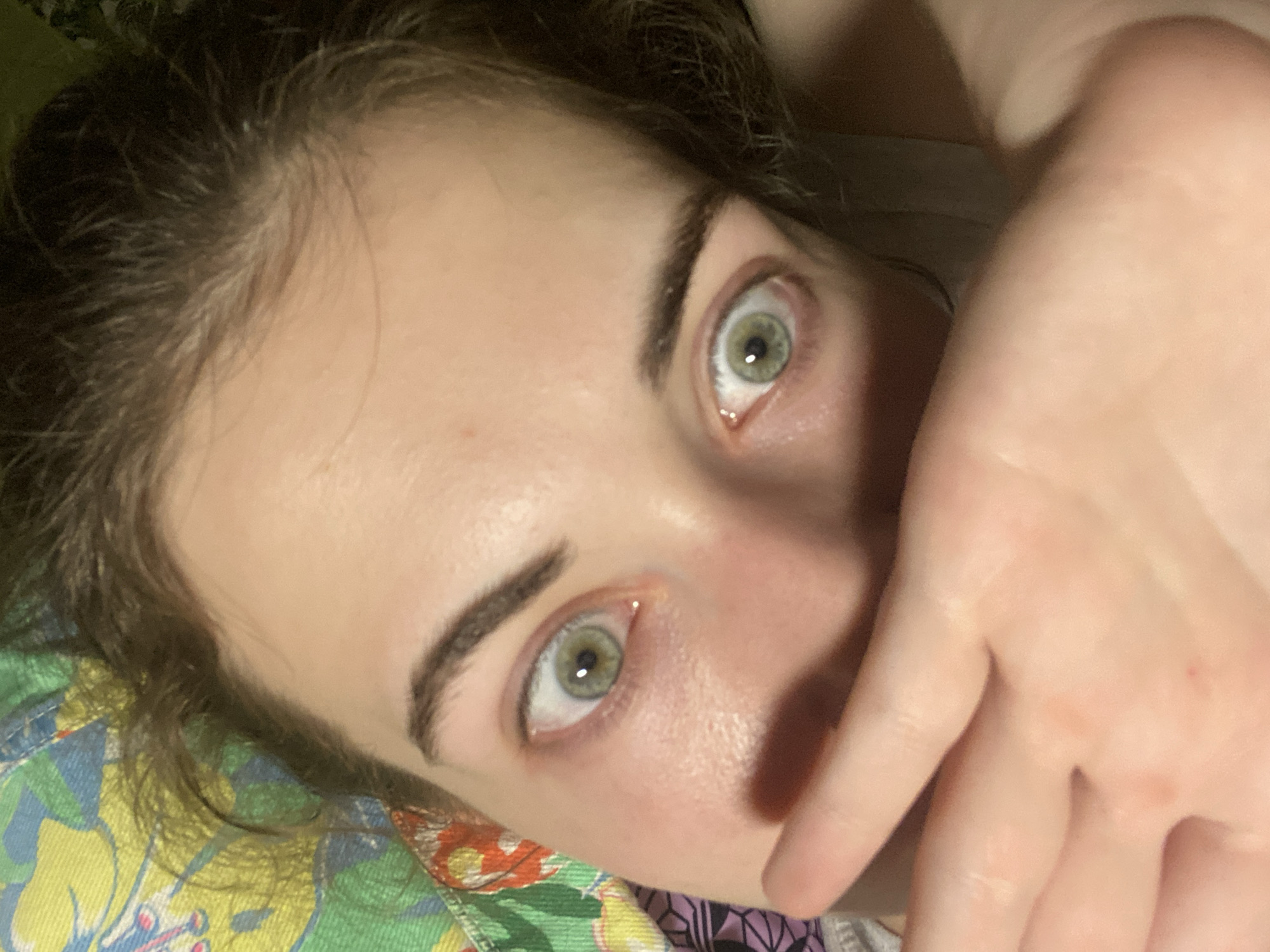 The image is a close-up of a person's face, focusing on their eyes and forehead, with their hand partially covering their mouth. The person has light skin and appears to have green eyes with a startled expression. Their eyebrows are dark and well-defined. The background is blurry but suggests a colorful, patterned fabric. The image was taken in Zavodoukovsk, Russia.