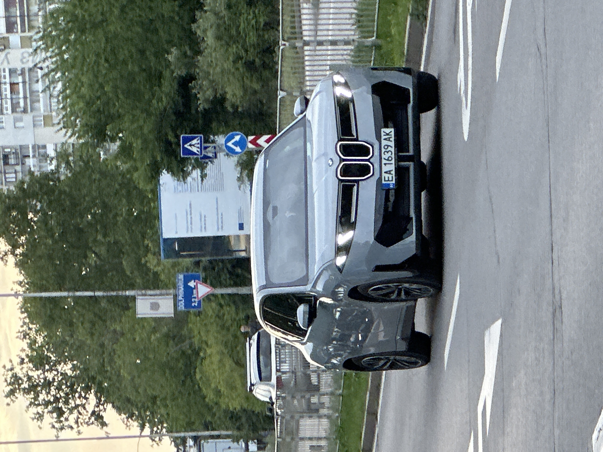 An image captured from a slightly elevated perspective shows a modern gray BMW iX SUV on a paved road in what appears to be an urban or suburban setting. The car is the primary subject, angled towards the viewer, revealing its distinctive large front grille with two prominent rectangular black sections and narrow headlights. Its license plate, "EA 1639 AK", is clearly visible on the front bumper, indicating a Bulgarian registration.
To the left of the car, a white vehicle is partially visible further down the road, behind a metal railing. The road features white painted markings. In the background, there are green trees and multi-story residential buildings. The lighting suggests it is daytime, possibly late afternoon, with soft, diffused light.
Several road signs are present. One blue rectangular sign in the mid-ground reads "DOLPHIN PROMENADE" and "2.3km". Nearby, a blue circular sign with a white arrow pointing right (mandatory right turn or direction) and a blue square sign with a white triangular emblem (indicating a pedestrian crossing ahead) are visible. Another sign, a white triangle with a red border, is also present, partially obscured. The image itself is oriented sideways, making the car appear to be driving vertically. The overall scene points to a location within Varna, Bulgaria, as per the license plate and contextual information. No people are visible.