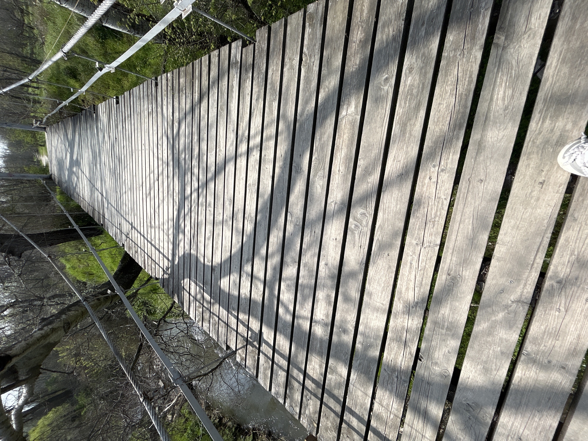 The image shows a wooden suspension bridge. The bridge has wooden planks laid parallel to each other with gaps between them. A wire rope runs along the side of the bridge, held up by metal posts. Below the bridge, vegetation and water can be seen. The light suggests a sunny day. The location is Plovdiv, Bulgaria.