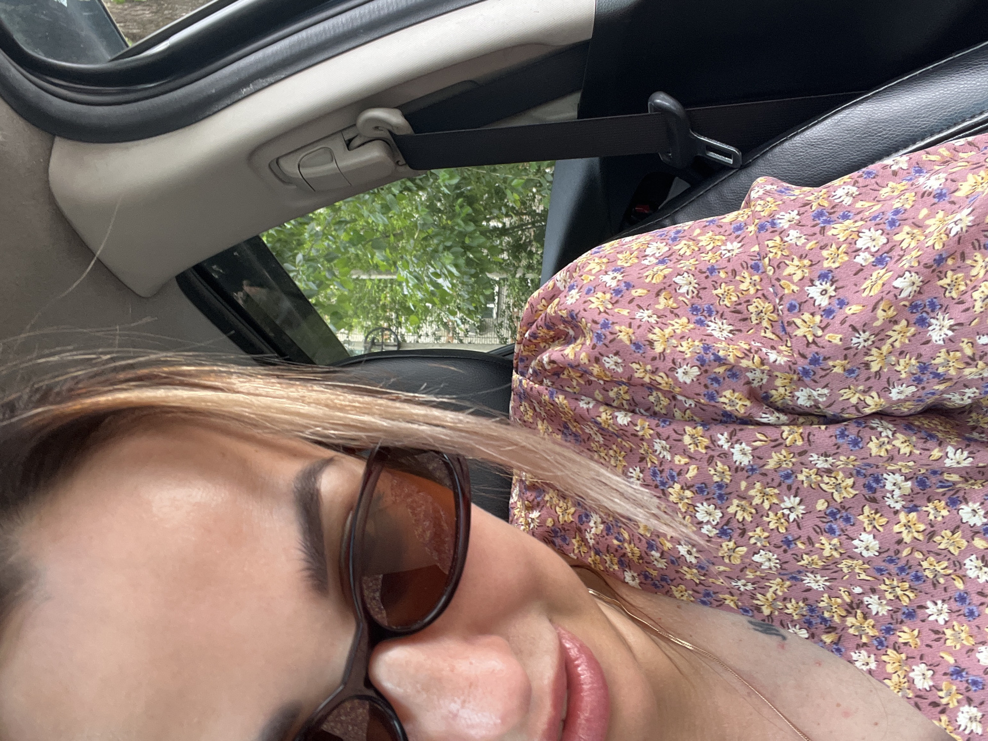 A woman is in the passenger seat of a car. She is wearing sunglasses and a floral dress. The car is parked in a location with trees and buildings visible outside the window. The seatbelt is visible.
