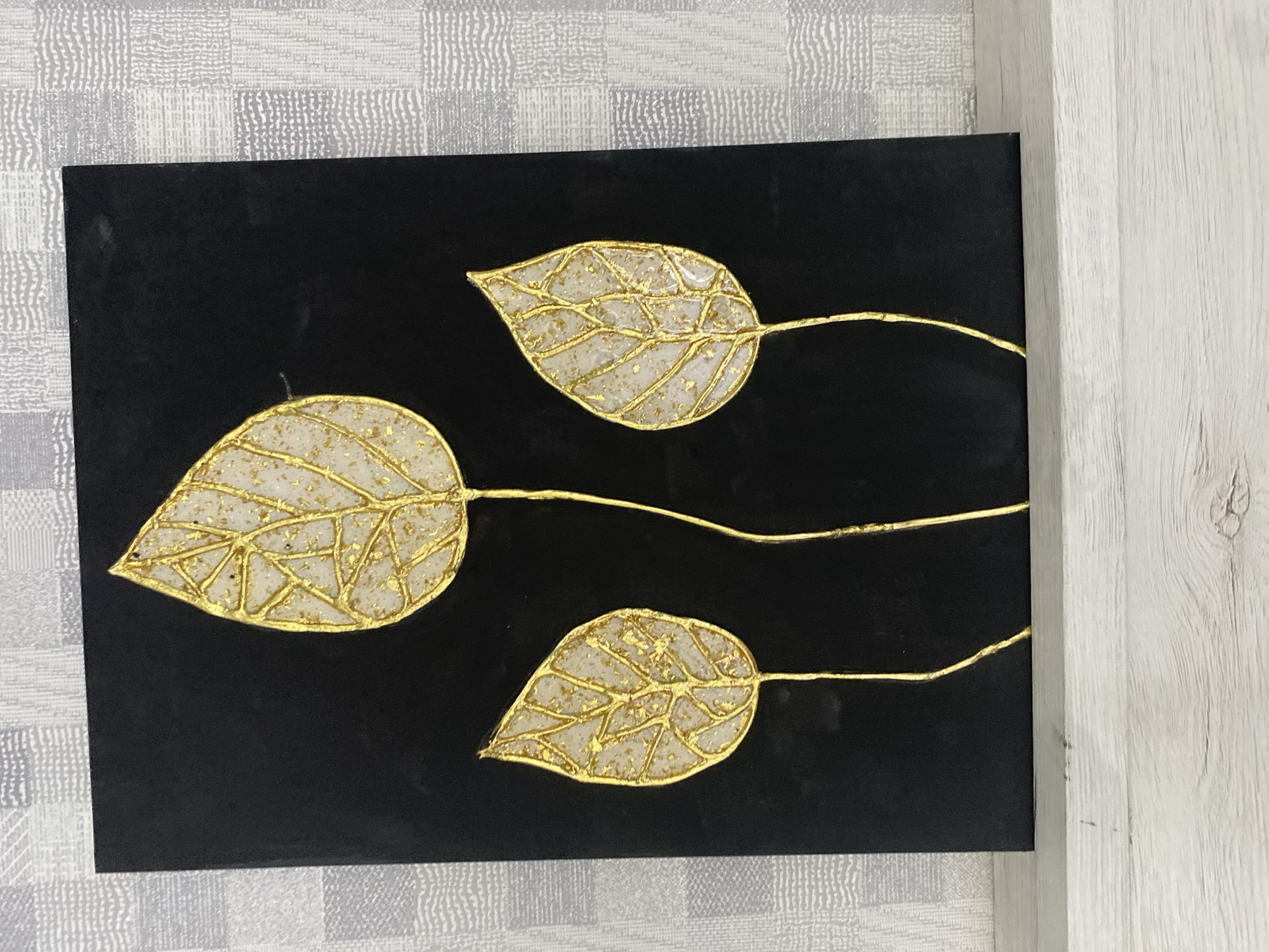 A black canvas features a minimalist design of three stylized leaves rendered in gold. The leaves, varying slightly in size and orientation, are outlined and veined with shimmering gold. The background of the canvas is a flat, deep black. The canvas is displayed in an indoor setting, leaning against a wall with a textured, grey and white patterned wallpaper. In the foreground, a light-colored, wood-grained surface is visible. The overall impression is one of elegant simplicity and metallic embellishment.