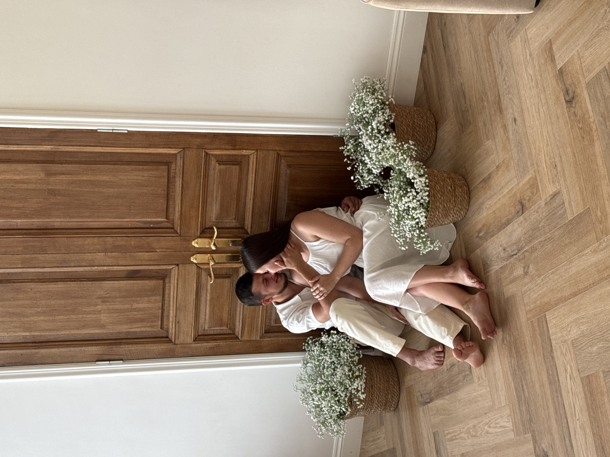 A man and a woman are sitting on the floor in front of a large wooden double door. The man is wearing a white short-sleeved shirt and cream-colored pants. The woman is wearing a white dress. They are facing each other and appear to be embracing. There are two woven baskets filled with small white flowers placed on the floor on either side of the couple. The floor is made of light brown wooden planks laid in a herringbone pattern. The wall behind them is painted white. The image was taken indoors. It is not possible to determine the specific location in the city of Izhevsk, Russia from the image alone.