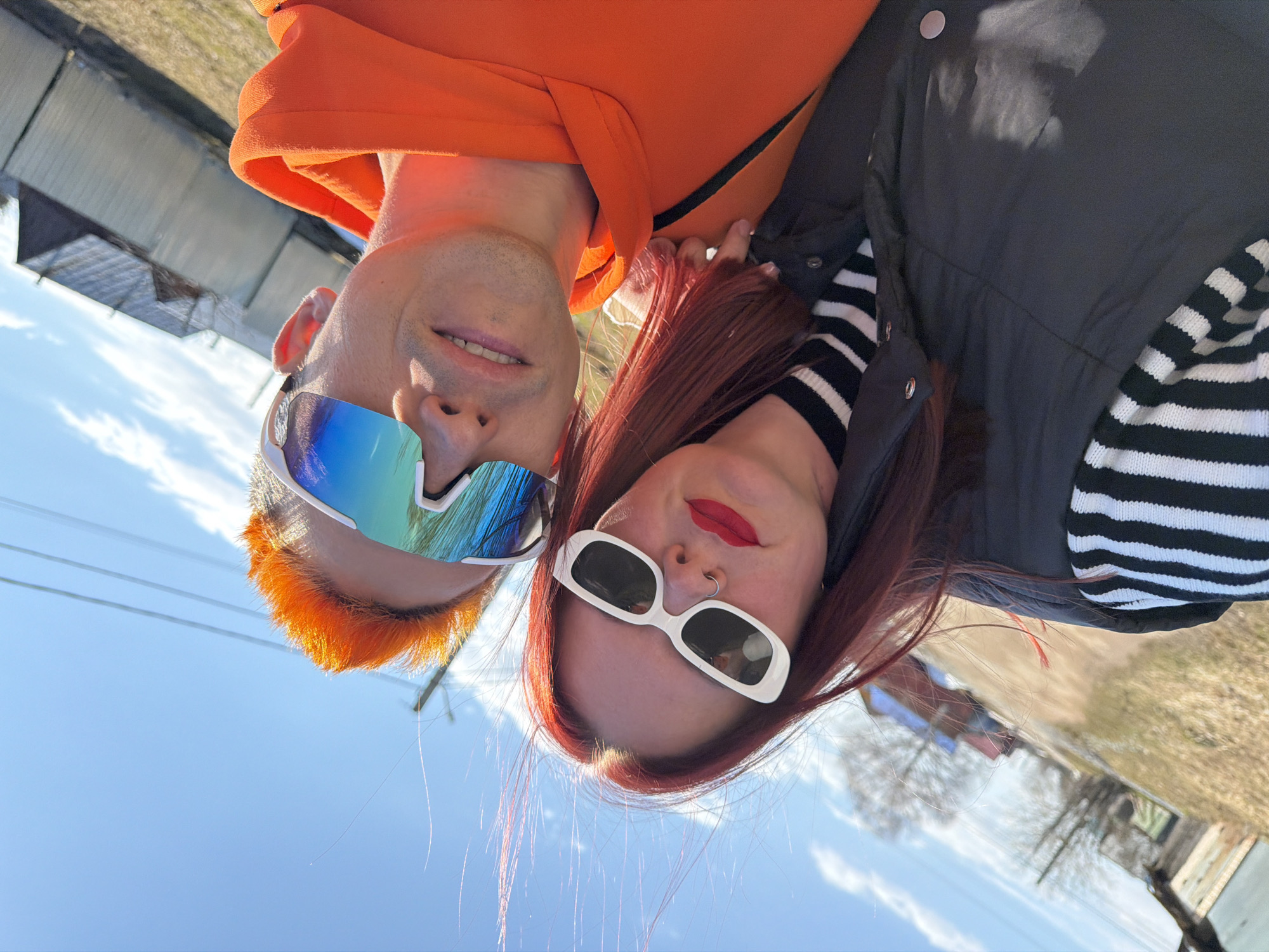 This media file captures a close-up selfie of two individuals, rotated 90 degrees counter-clockwise, portraying them from a low angle against a bright sky. The subjects appear upside down relative to the background.
On the left, a man with vibrant, bright orange hair and stubble on his face smiles gently towards the camera. He wears a bright orange hooded sweatshirt and large, futuristic-style reflective sunglasses with blue and green tints. His mouth is slightly open, showing part of his teeth.
On the right, a woman with long, rich red hair frames her face. She wears fashionable white, rectangular sunglasses that obscure her eyes. Her lips are painted with vivid red lipstick, and she has a small nose ring on her left nostril. She smiles slightly. Underneath a dark, possibly black, puffer vest or jacket, a black and white striped long-sleeve top is visible. Her red hair catches the sunlight, showing strands illuminated against the sky.
The activity is a shared selfie, with both subjects looking towards the camera, seemingly happy or content.
The background reveals a clear, bright blue sky with sparse white clouds, suggesting a sunny day. In the distance, bare trees, characteristic of late winter or early spring, are visible. Utility lines (likely power lines) stretch across the upper portion of the sky. Faintly visible structures, possibly houses or sheds, and a patch of dry, light-brown ground suggest an outdoor, possibly rural or semi-urban, setting in Chaikovsky, Russia. There is no visible text in the image.
