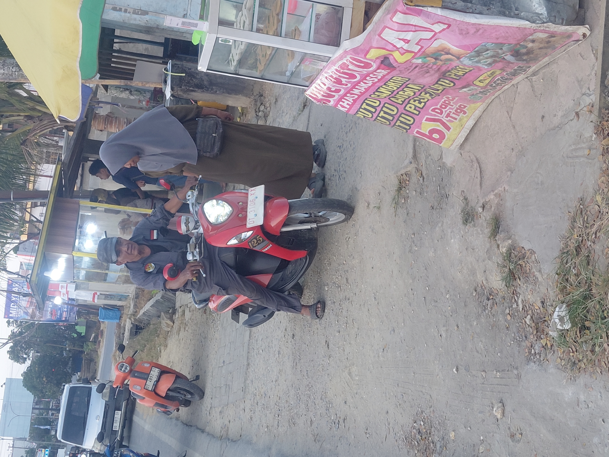 This is a medium-shot, eye-level, outdoor photograph taken during the day in Kendari, Indonesia. The scene depicts a man in a dark gray uniform and a dark gray cap riding a red scooter. He is looking towards the right side of the frame. Next to him, on his right, a woman wearing a grey hijab and a dark green long-sleeved garment stands. She is looking towards the left and has a black bag slung across her shoulder. The scooter the man is riding is red with white accents and has a license plate that reads "DT 3106 XX". Another orange scooter is parked in the background to the left, with its license plate reading "DT 4892 01". A white van is visible behind the orange scooter. The environment is a dusty, unpaved roadside with some sparse vegetation. A pink banner with Indonesian text and images of food is partially visible on the right side of the frame, leaning against a glass display case, suggesting a street-side food stall. The sky is visible at the top of the image and appears to be overcast or hazy. The overall impression is a typical street scene in a provincial city.