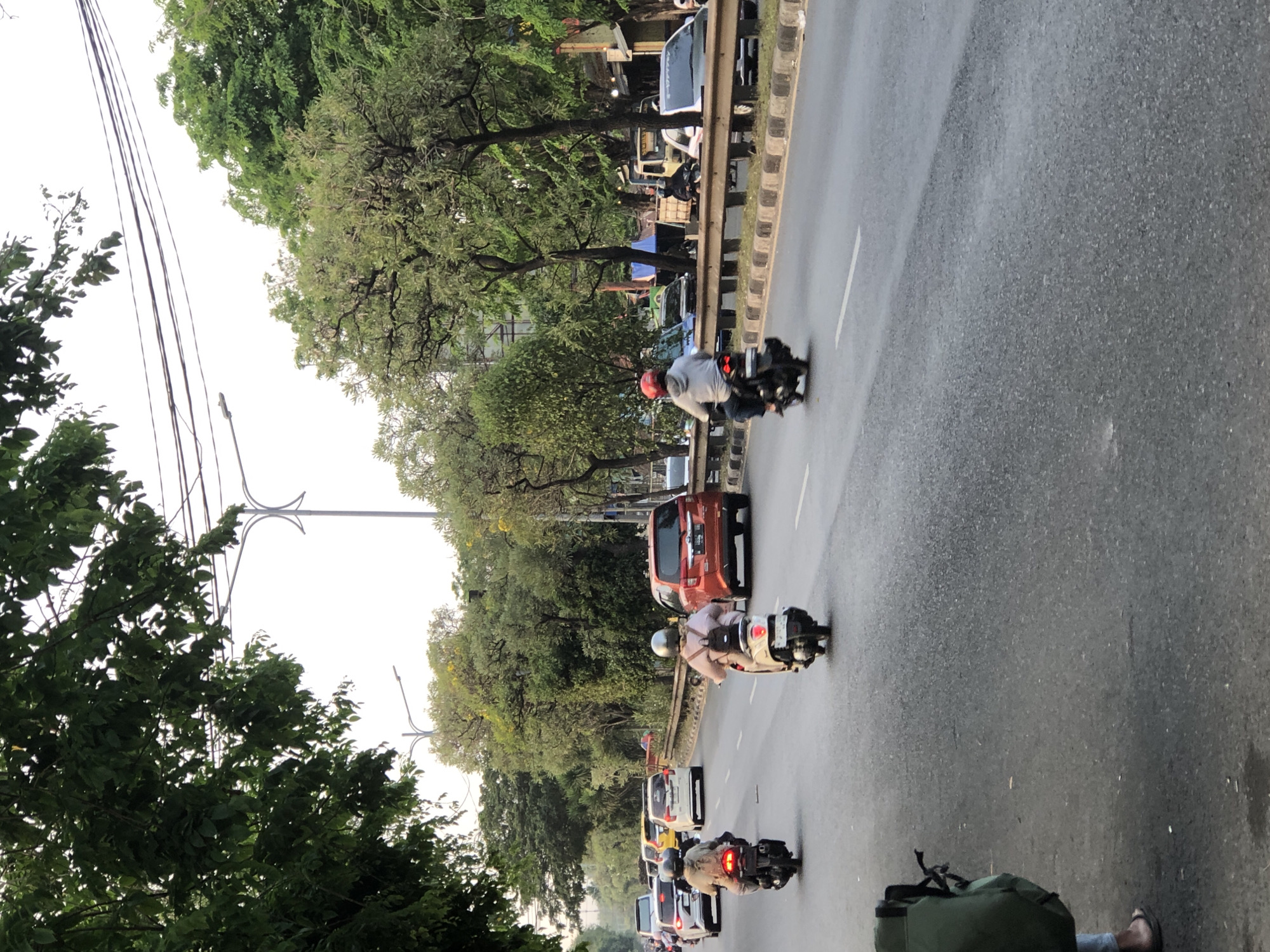 The image depicts a street scene in Kludan, Indonesia, likely during daylight hours. The road is paved and appears to be a multi-lane thoroughfare with traffic. In the foreground, a person is partially visible on the left, wearing sandals and carrying a large green backpack.
Several motorcycles are in motion on the road. Two motorcycles are closer to the camera, each with a rider wearing a helmet and a backpack. Further down the road, other vehicles, including cars and at least one orange SUV, are visible in traffic. Trees line the side of the road, with some overhead wires and streetlights also present. The sky appears overcast or hazy. The overall impression is of a typical urban or suburban street with moderate traffic flow.