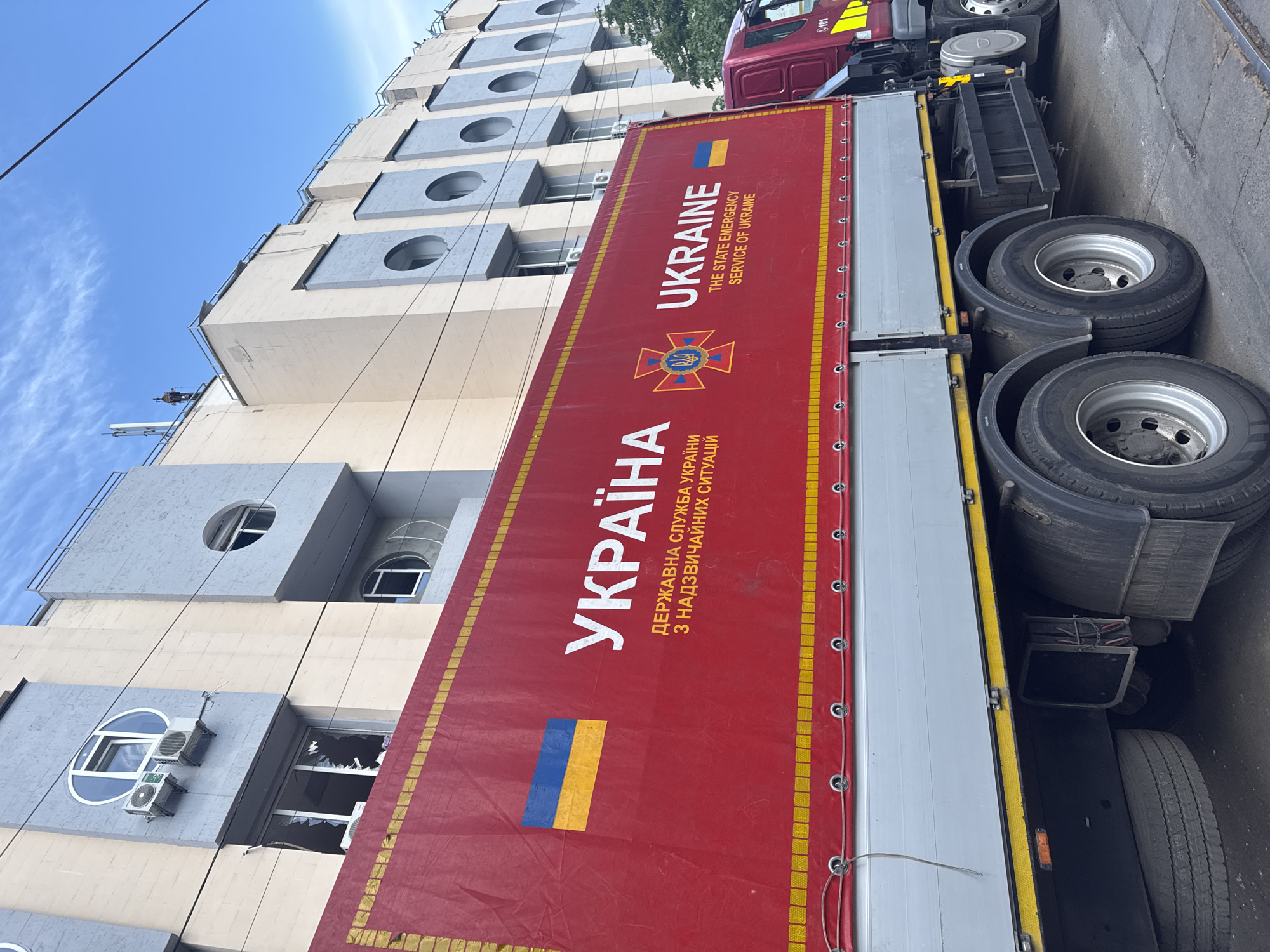 The image shows a truck, likely a utility or service vehicle, parked on a street in Kyiv, Ukraine. The side of the truck is emblazoned with the word "УКРАЇНА" (Ukraine) in large letters, along with the Ukrainian flag and the emblem of the State Emergency Service of Ukraine. The building next to the truck has several stories with round windows and a window that is broken out. Power lines are present. The overall scene indicates a setting that has been impacted by conflict or damage.