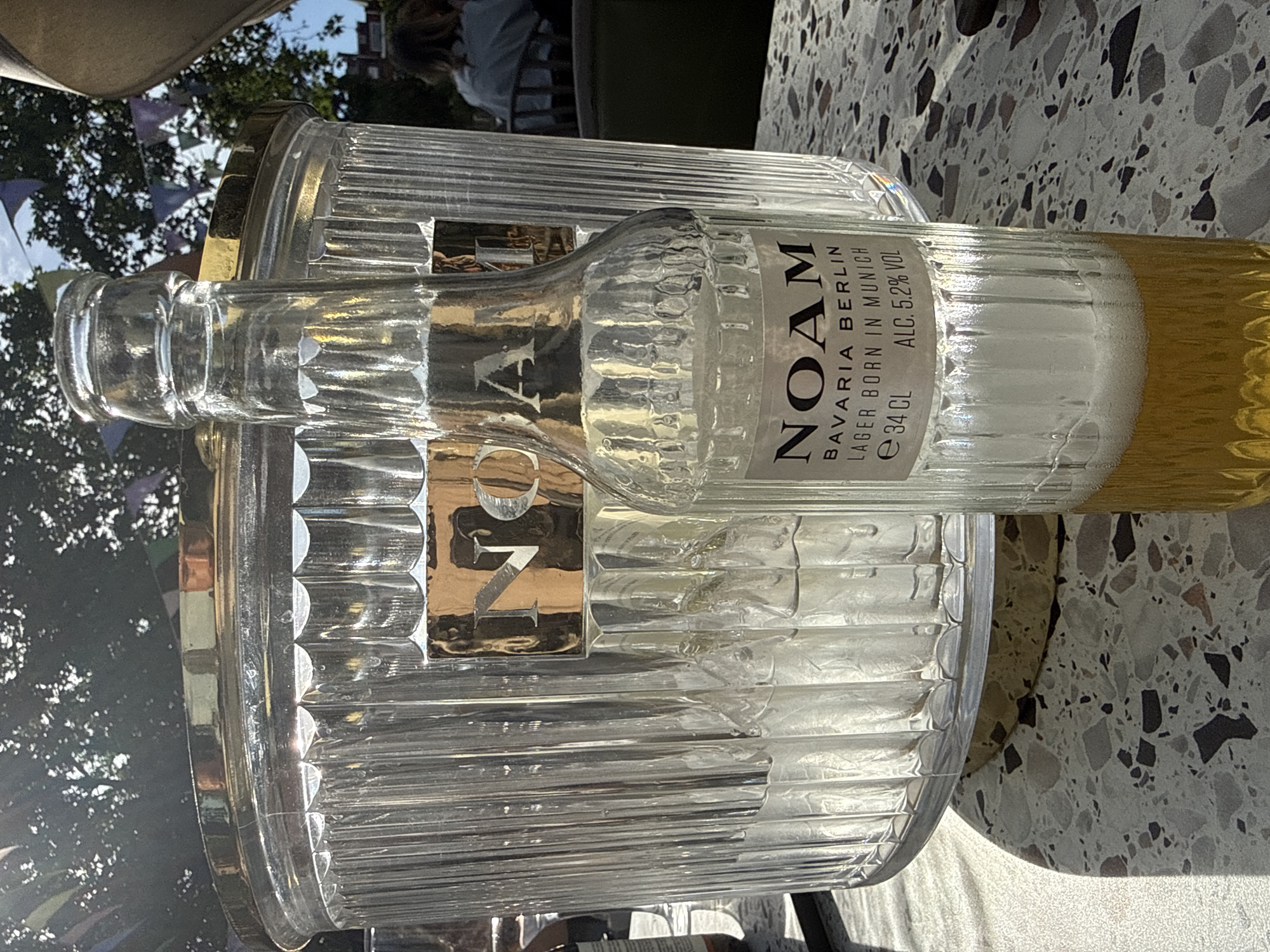 An eye-level, angled shot captures a clear glass bottle of "NOAM BAVARIA BERLIN LAGER BORN IN MUNICH" resting on a translucent, circular, ridged ice bucket. The bottle is partially filled with a golden liquid. The scene unfolds on an outdoor terrazzo-like surface. Background elements include a person seated in a chair, greenery, and building facades, suggesting an urban environment. A glimpse of blue pennant flags hangs from trees. This setting is consistent with an outdoor space in the city of London, United Kingdom.