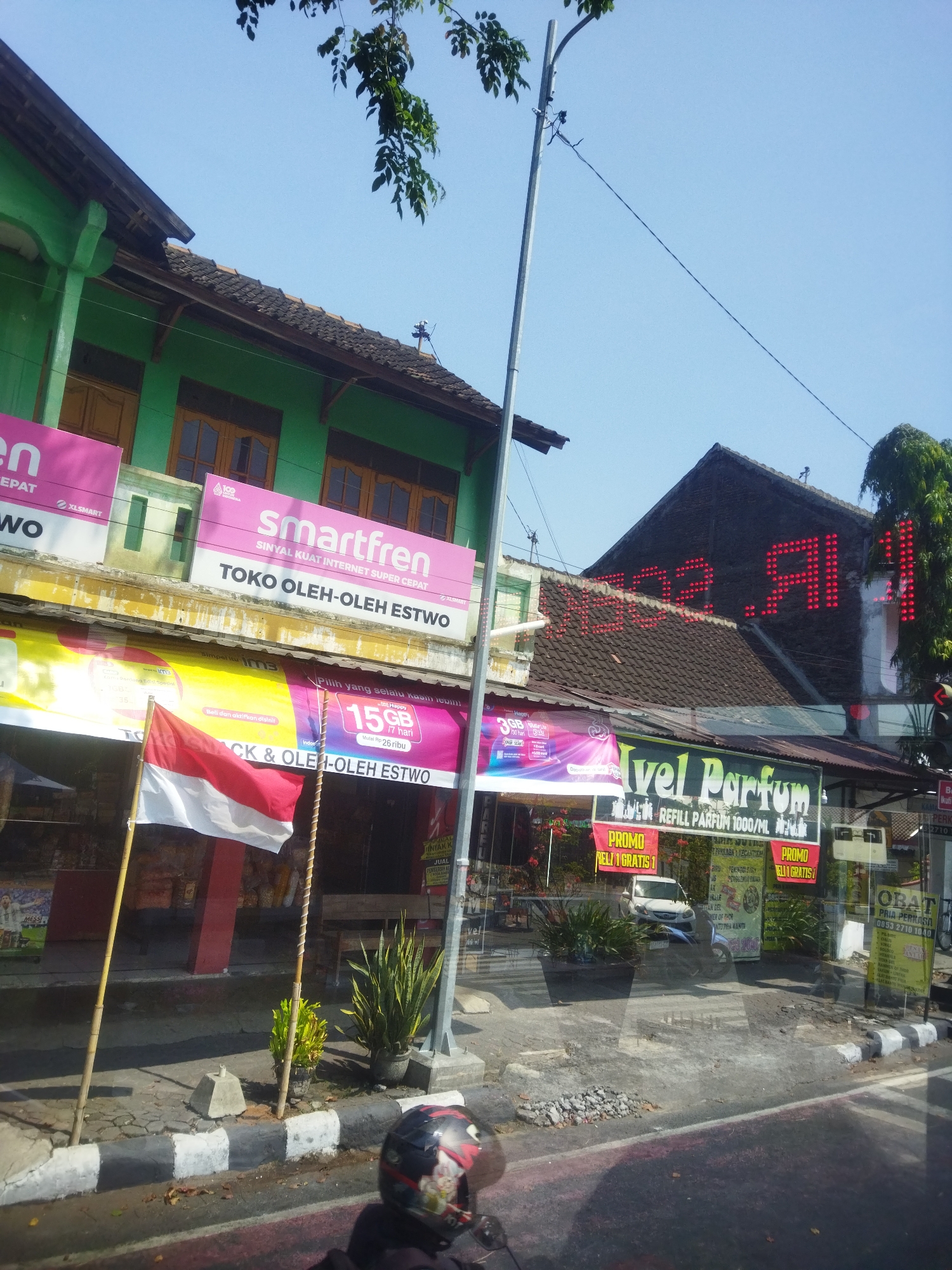 A medium shot captures a street scene in Sukoharjo, Indonesia, on a bright, clear day. The foreground features the shoulder of a road, with a concrete curb painted with alternating black and white stripes. A motorcycle helmet, decorated with a cartoon character and flames, rests on the road surface, partially obscuring the curb. To the left, an Indonesian flag, red and white, stands upright on a bamboo pole.
Behind the flag, a row of shops lines the street. The buildings have green and brown facades, with visible windows and doorways. Prominent signage adorns the shops, including a large pink banner for "smartfren" with the tagline "SINYAL KUAT INTERNET SUPER CEPAT" and "TOKO OLEH-OLEH ESTWO." Below this, a yellow banner displays "im3" and "15GB". Another shop features a sign for "Nivel Parfum" with "REFILL PARFUM 1000/ML". Red LED lights spell out "MAZA.AI" on the dark roof of a building.
Various potted plants, including a snake plant, are strategically placed along the storefronts, adding a touch of greenery to the urban environment. A white car is parked in front of one of the shops. The overall impression is one of a bustling commercial area on a typical Indonesian street.