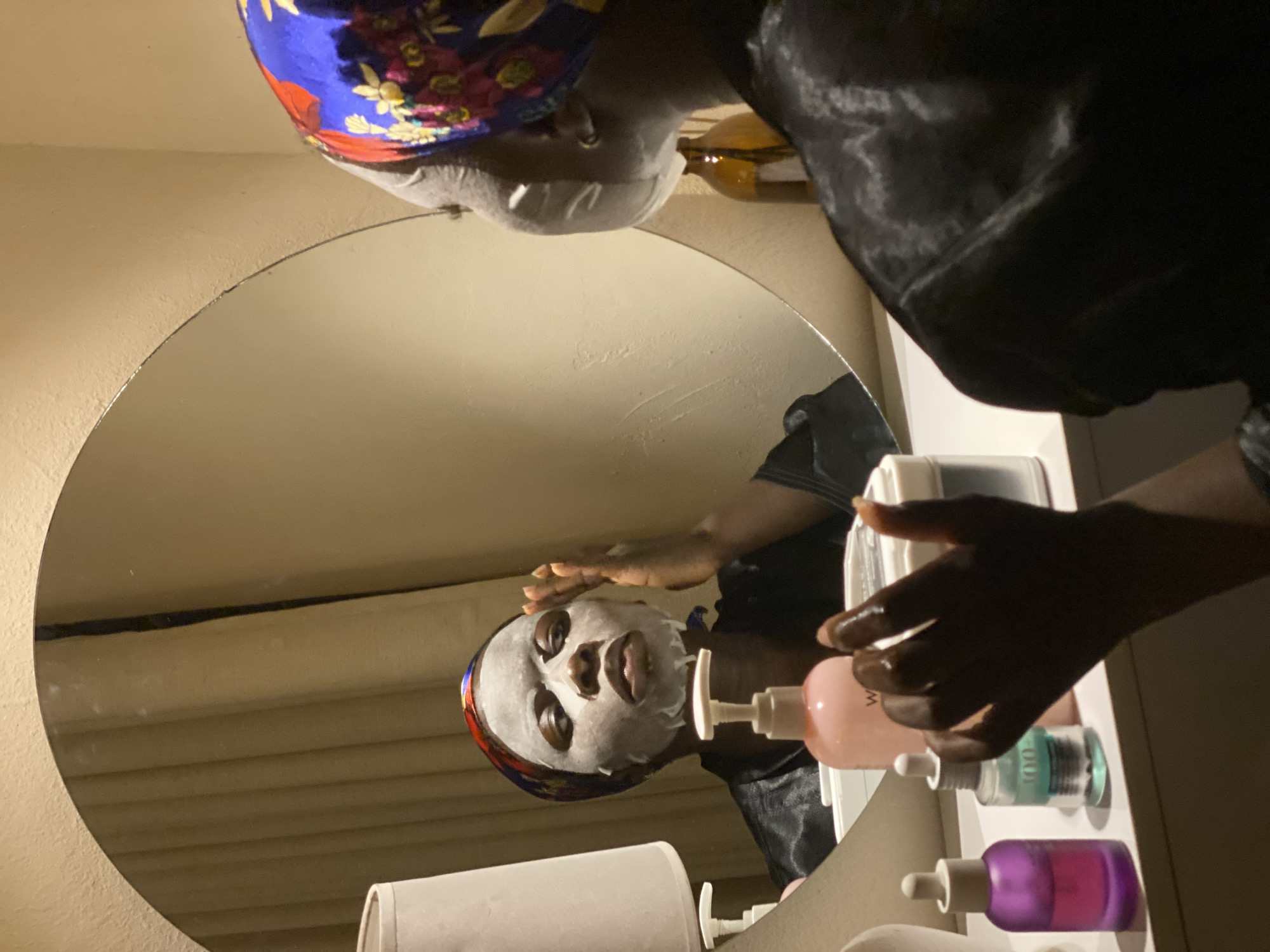 A person with a white facial mask is visible in a round mirror, presumably in an indoor setting. The person is wearing a blue and floral patterned head covering and a dark silky robe. Their right hand is raised near their face, seemingly adjusting the mask. Several skincare bottles of different colors (pink, turquoise, purple) are placed on a surface near the mirror. The background appears to be a plain wall, possibly in a bathroom or bedroom. There is no indication in the image that it is in Jos, Nigeria, or any location in particular.