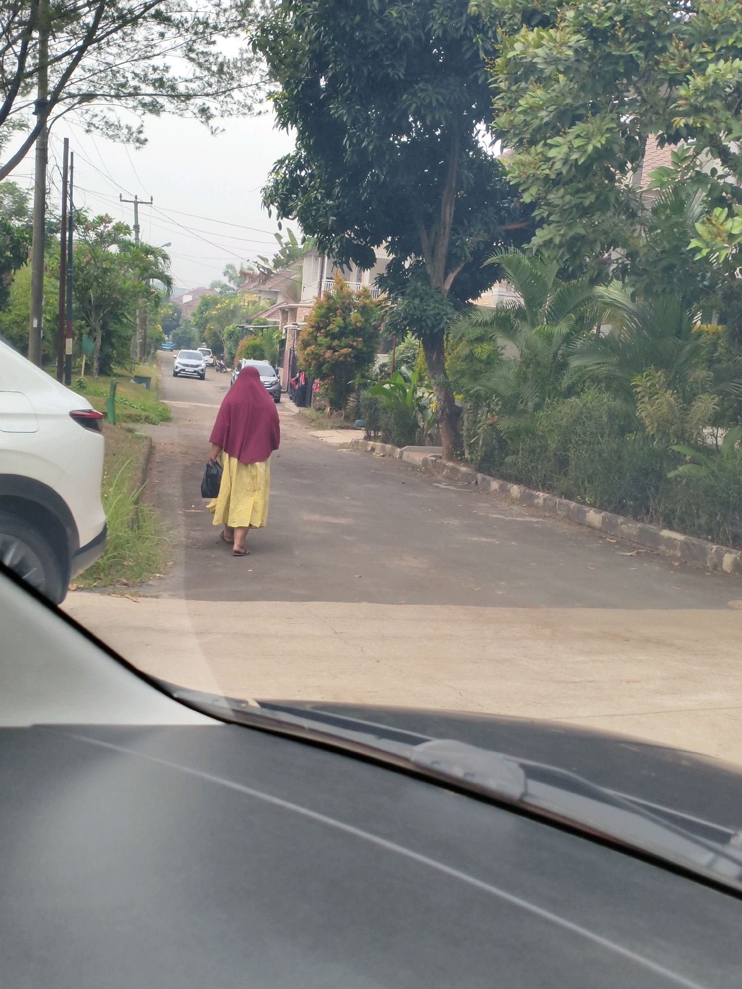 A woman wearing a maroon hijab and a yellow dress walks along a paved road in Sukaraja, Indonesia. She carries a black bag in her left hand. Several cars are parked and moving on the road, which is bordered by lush green trees and houses. The scene is captured from the perspective of someone inside a vehicle, as the car's windshield and hood are visible in the foreground. The sky is overcast.