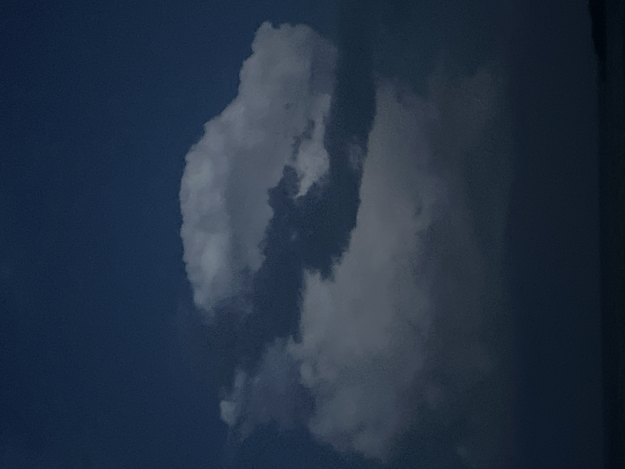 The image depicts a dark, cloudy sky over a body of water, likely at dusk or dawn. A large, white cloud formation dominates the upper half of the image, with darker clouds interspersed. The lower portion of the image shows a dark, indistinct horizon line that suggests land or water. The scene is devoid of any human presence or man-made structures. This image does not depict Novopichugovo, Russia.