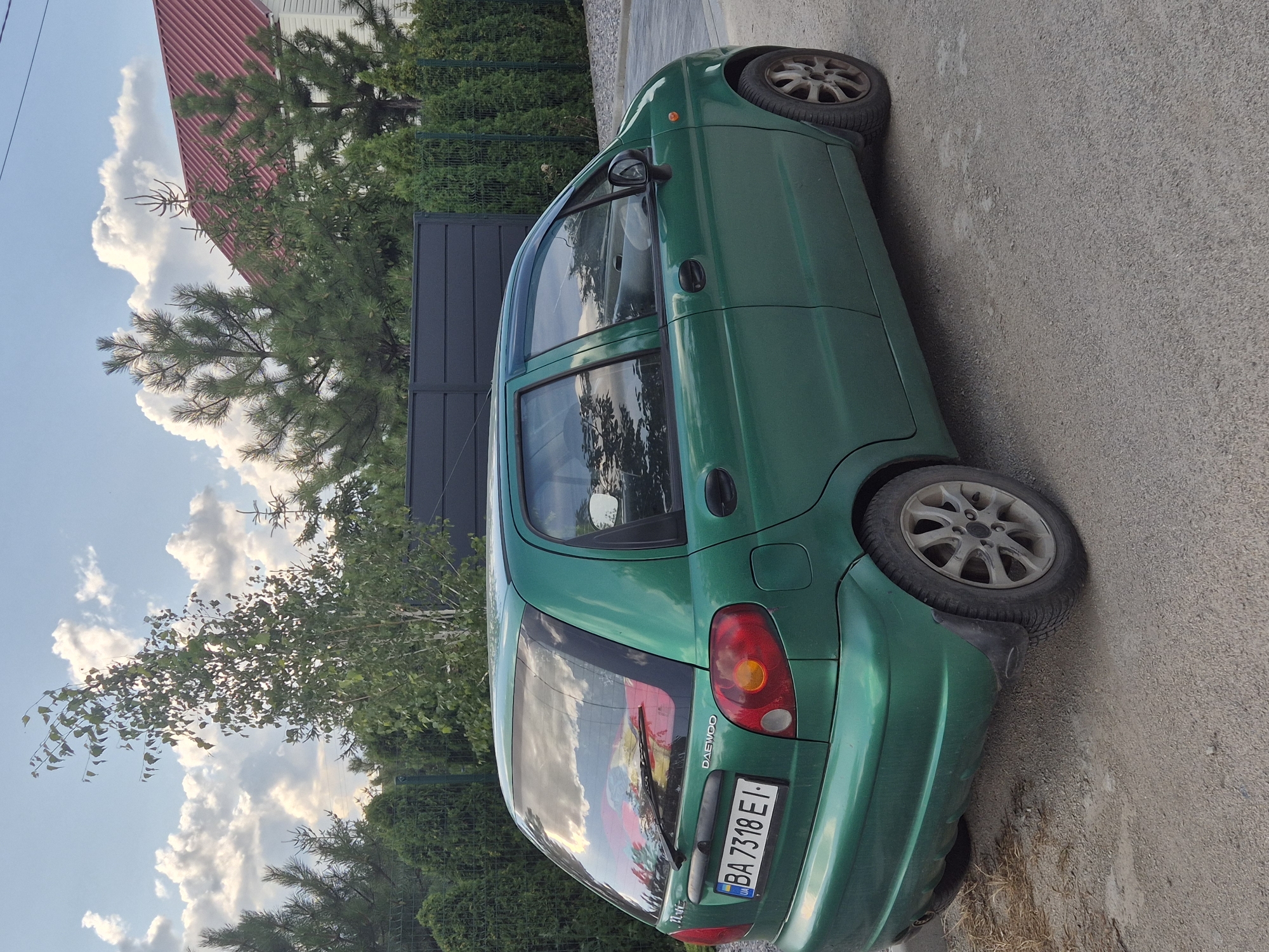 A green Daewoo Matiz car is parked on a gravel surface in front of a dark metal fence and a row of evergreen trees. Behind the fence, a white house with a red roof is visible, surrounded by trees. The sky is partly cloudy with large, white cumulus clouds. The car has a Ukrainian license plate reading "BA 7318 EI". The overall scene depicts a residential outdoor setting in Sozonivka, Ukraine.