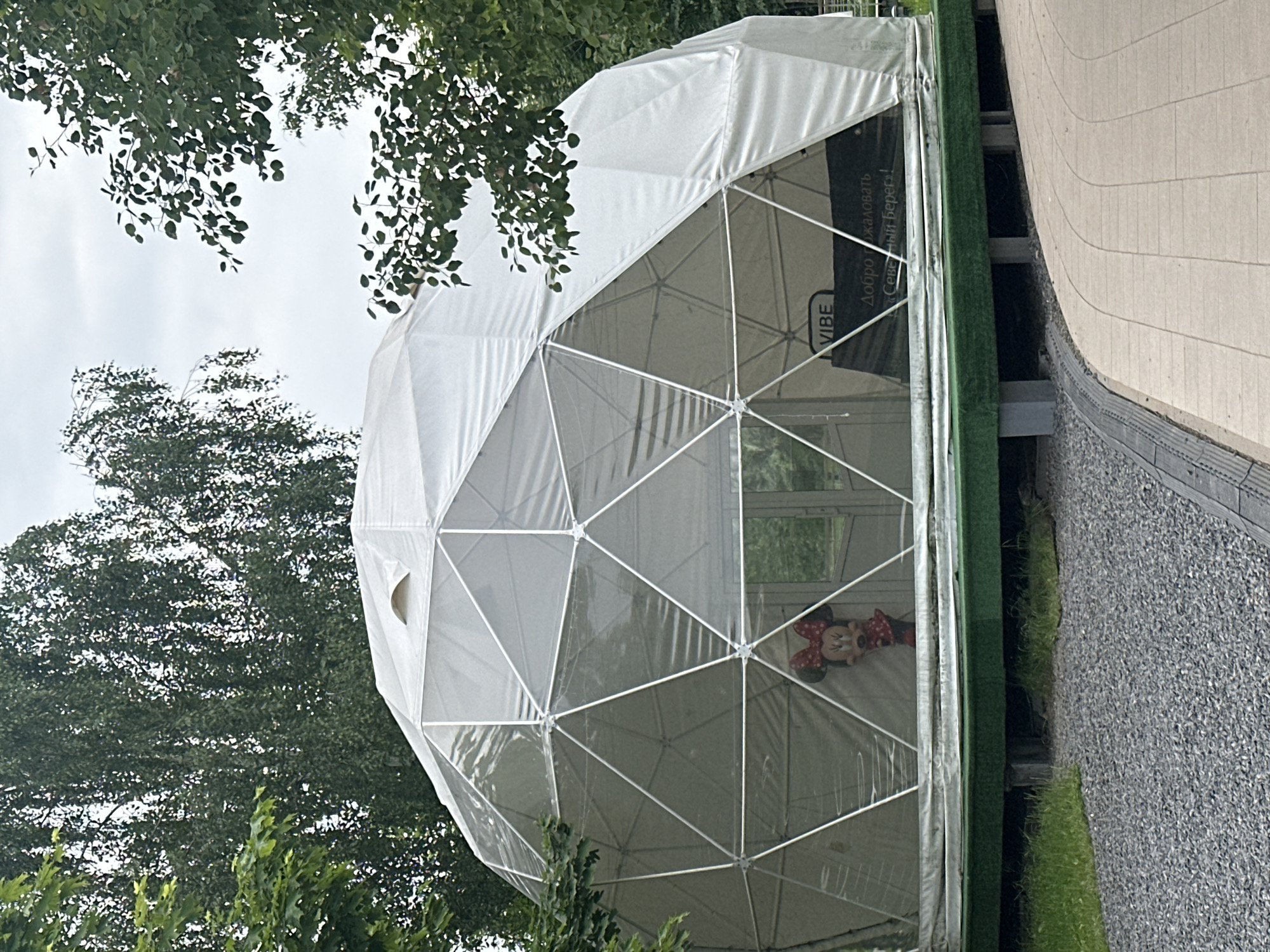 A geodesic dome tent is situated outdoors, possibly in a park or recreational area. The tent is white and made of a fabric material stretched over a metal frame, creating a geometric pattern of triangular panels. Inside the tent, a Minnie Mouse balloon is visible, appearing to be a decoration. A sign with text in Cyrillic script, which translates to "Welcome to 'Sunny Beach'!", is also present within the tent. The tent is elevated on a platform covered with artificial green turf, and the ground around it consists of gravel and a paved walkway. Trees with green foliage surround the structure, and the sky is overcast. The scene appears to be in the city of Minsk, Belarus, likely at a location known as "Sunny Beach".