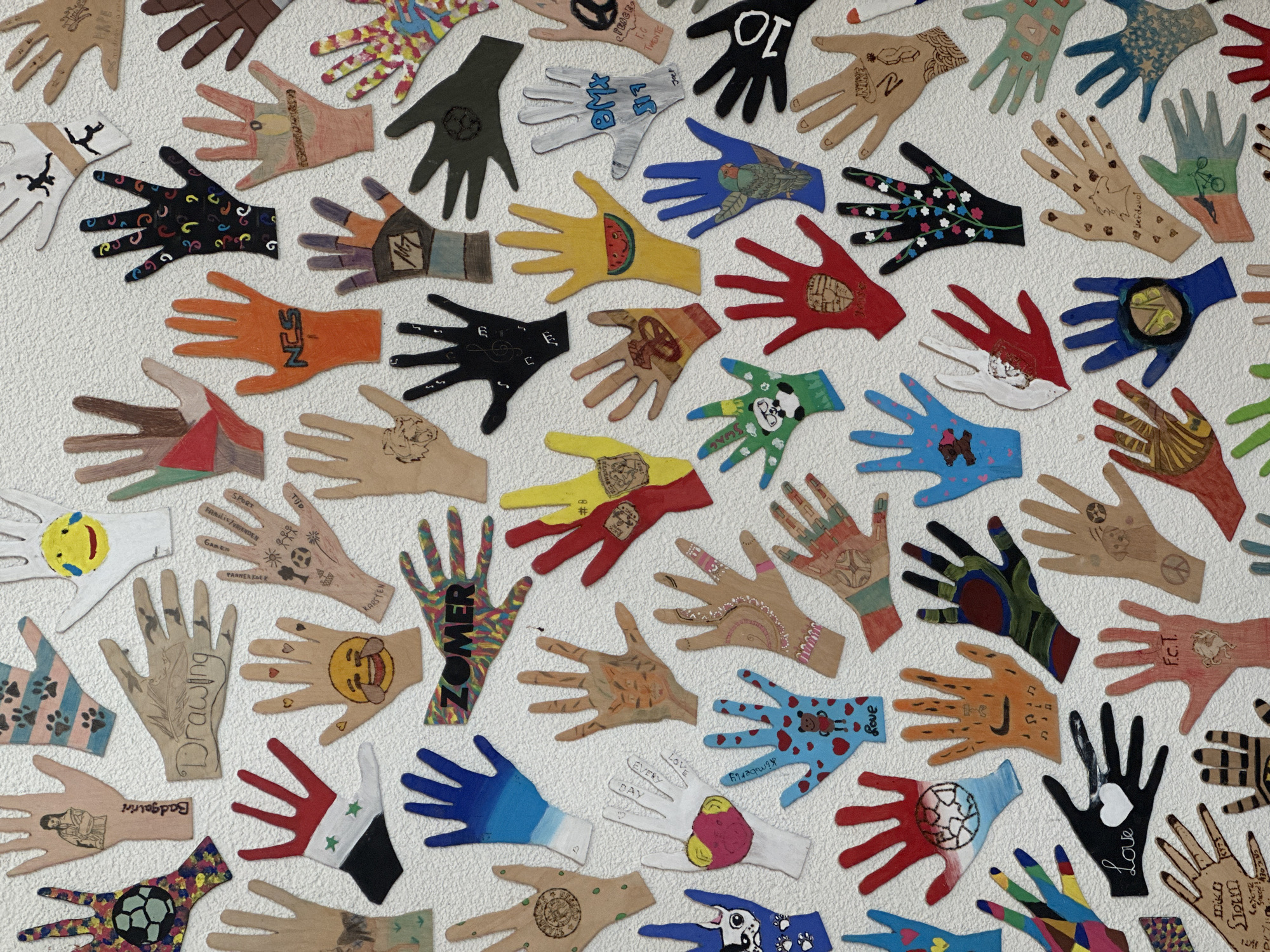 The image displays a collection of diverse handprints, each uniquely decorated and adhered to a textured white wall. The handprints vary in color, size, and artistic embellishment, featuring drawings, text, and patterns. Some handprints depict animals, while others have symbols, flags, or abstract designs. One handprint, in the lower left quadrant, displays the text "Love" with a heart. Another handprint, located to the right and slightly below the "Love" handprint, contains the text "F.C.T." above a drawing of a creature. The collection appears to be a collaborative art project, possibly created by children or a community group. The wall's textured surface suggests it might be an interior wall with a stucco or similar finish. The lighting is even, indicating a well-lit indoor environment. No people are visible in the image. The overall impression is one of creativity and personal expression through art. This artwork is located in the city of Borne, Netherlands, specifically at a sports club or community center.