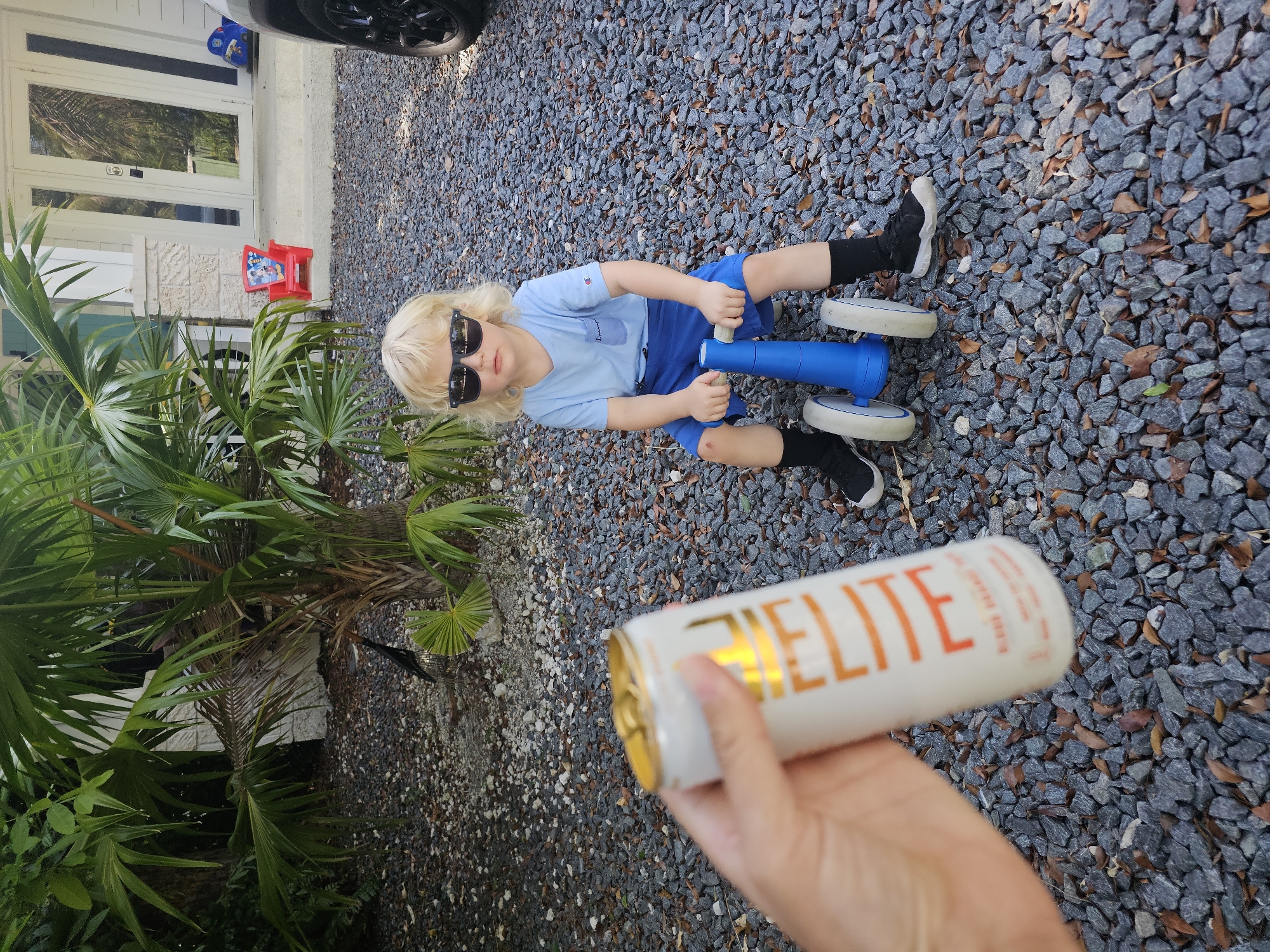 A young boy with blonde hair, wearing sunglasses, a light blue t-shirt, blue shorts, and black socks, sits on a gravelly surface. He is holding the handlebars of a blue and white toy vehicle with three wheels. To the right, a hand is holding up a white can with orange text. The can has a gold-colored top. The background shows a house with white doors and stone accents, and lush green palm fronds. The gravelly surface is interspersed with dry leaves, suggesting an outdoor setting. The time of day appears to be daytime, possibly late afternoon, with shadows visible on the ground. The scene is set in Islamorada, USA, a location known for its tropical climate. The boy's posture and sunglasses suggest a casual, relaxed atmosphere. The can has visible text that appears to read "ELITE" in large orange letters.