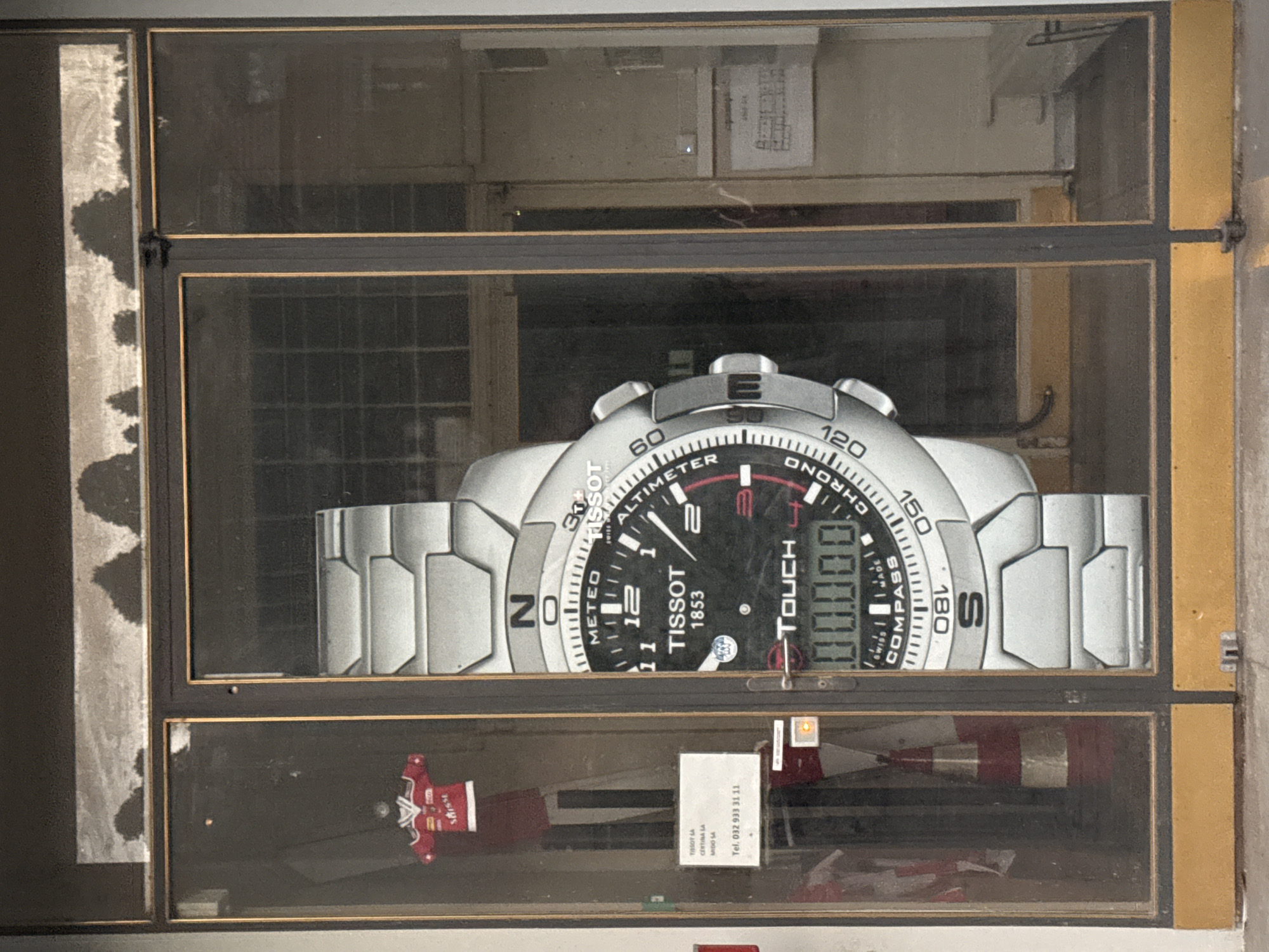 An outdoor, eye-level, medium shot captures a large Tissot watch advertisement displayed behind a glass storefront. The advertisement is positioned centrally on the right side of the frame, with the watch face angled slightly towards the viewer. The watch is silver with a black face featuring numerous markings and a digital display showing "00.00.00". The Tissot logo and "1853" are visible on the watch face. To the left of the watch advertisement, through the glass, a red and white traffic cone is visible, along with a small red and white shirt hanging on a mannequin or display. A white sign with text and a telephone number is also present on the left. The storefront has dark frames and the ground appears to be concrete with some yellow markings. The image is taken in Le Locle, Switzerland, a city known for its watchmaking industry.