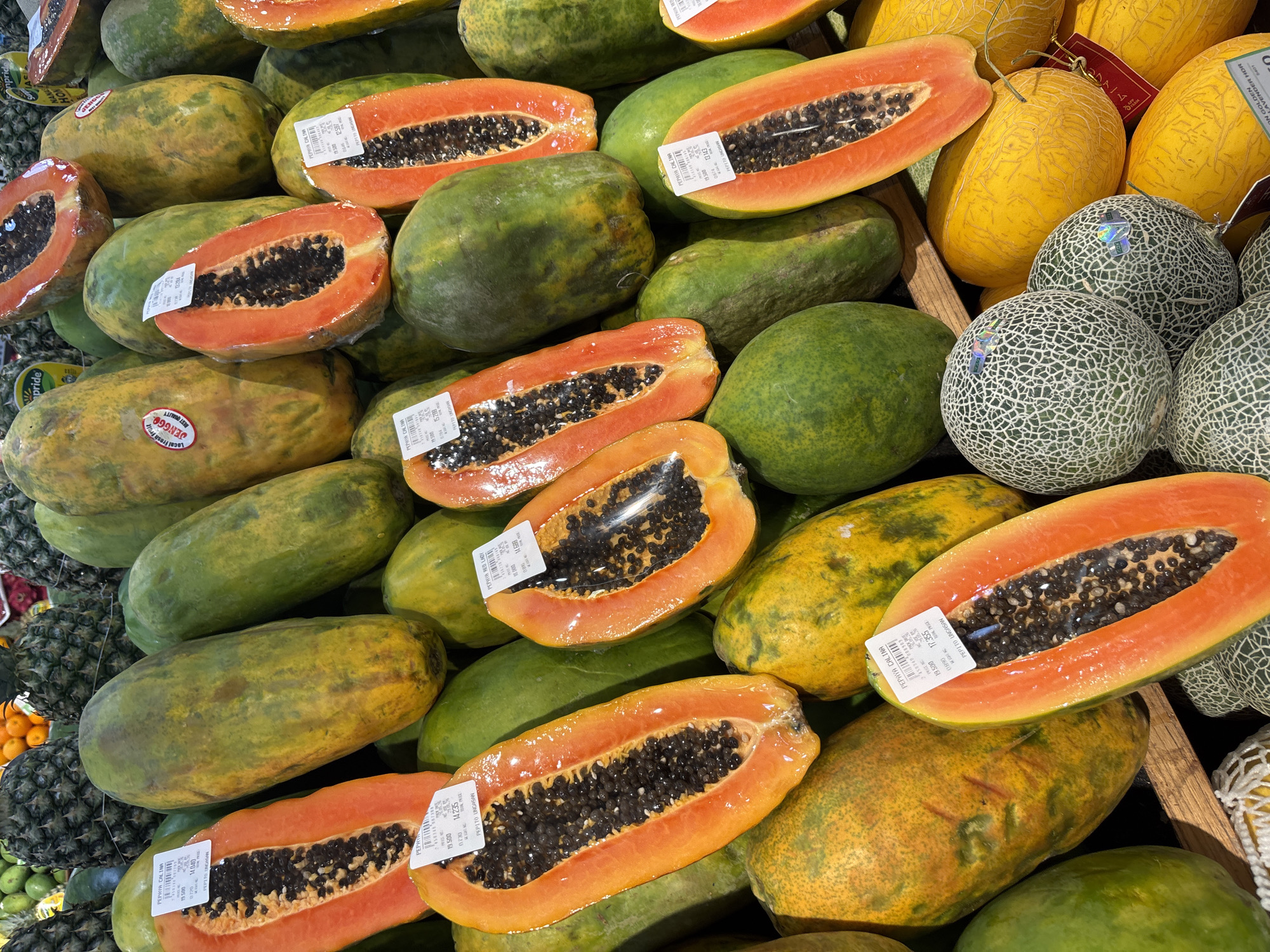 The image depicts a vibrant display of fruits at a store in Kuta Selatan, Indonesia, identified by "PEPITO UNGASAN" labels. The main subject is a variety of halved papayas, showing orange flesh and black seeds, some covered in plastic wrap with pricing stickers. There are also whole green papayas with yellow spots and green netted melons, and yellow oval melons visible. At the left of the image, pineapples are seen. The fruits are packed together, suggesting a market or grocery store setting. The pricing labels have Indonesian text.