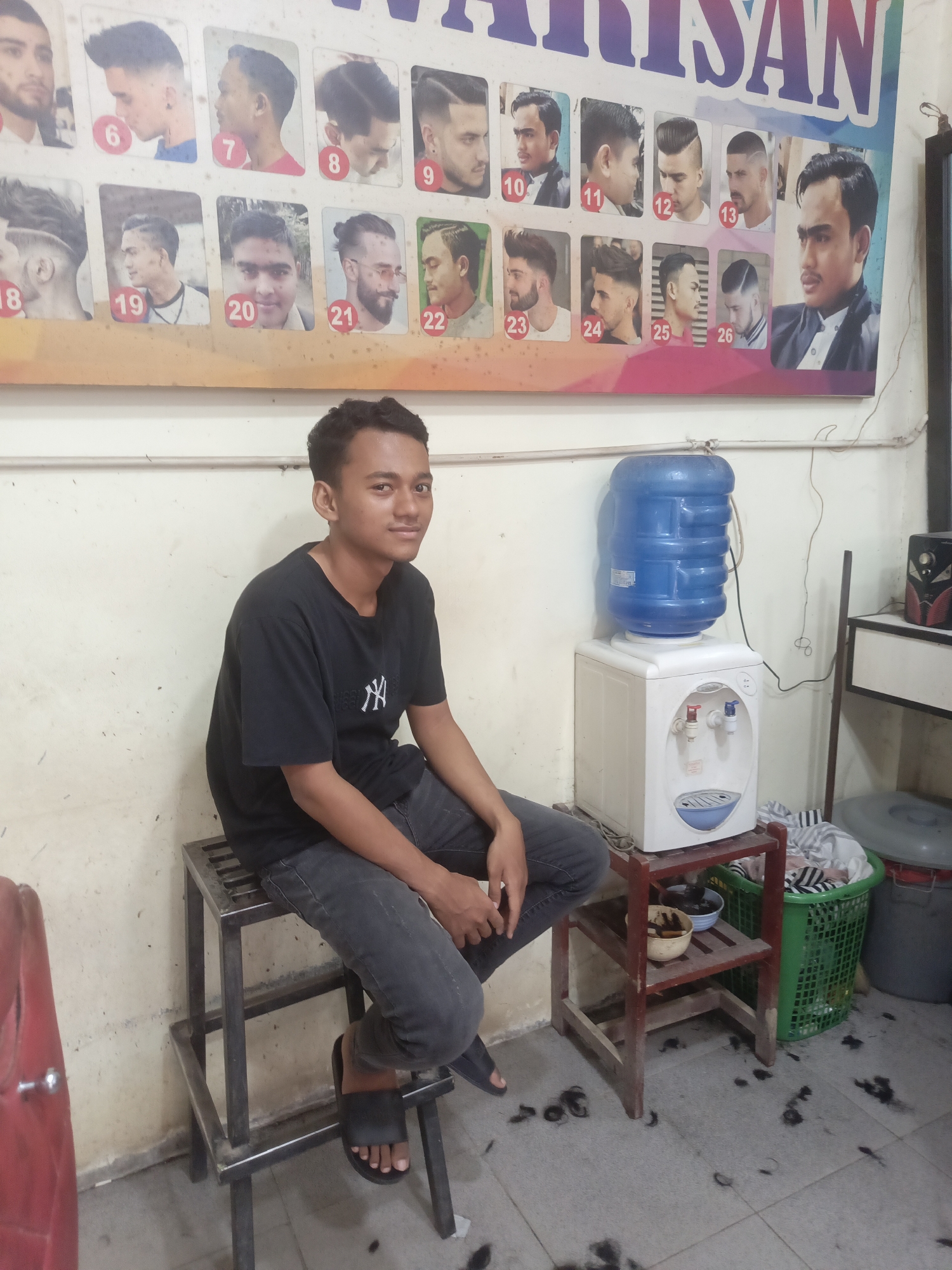 The image depicts the interior of a barbershop, likely located in Lhokseumawe, Indonesia, as indicated by the text on the wall. A young male individual, appearing to be in his late teens or early twenties, is seated on a metal stool in the foreground. He is wearing a black t-shirt with a white logo and dark grey jeans. He is looking towards the camera with a slight smile. Behind him, a white water dispenser with a blue top is prominently featured. To the right of the dispenser, a wooden shelf holds two bowls, one with a beige color and the other with a darker color, and a green laundry basket filled with clothes. The floor is tiled and scattered with hair clippings, indicating recent activity. On the wall behind the young man is a poster displaying various hairstyles, each numbered from 6 to 26, suggesting these are examples of haircuts offered at the establishment. The overall lighting suggests an indoor environment, possibly illuminated by artificial light.
